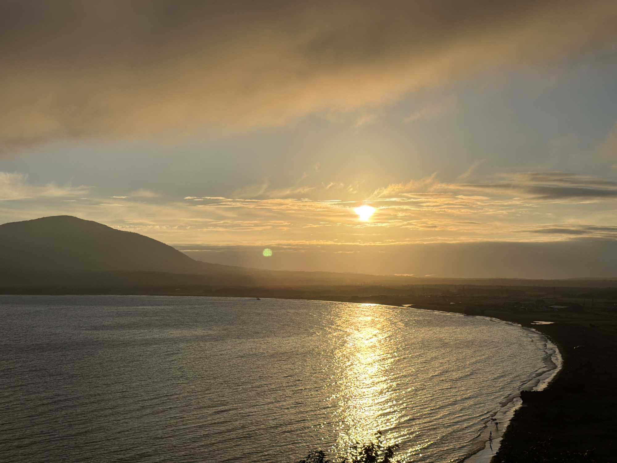 The image captures a panoramic view of a coastal landscape at sunset. A large, dark mountain range dominates the left side of the frame, its silhouette softened by the golden light. The ocean occupies the majority of the foreground, with its surface shimmering under the sun's reflection. The water exhibits a gentle ripple, suggesting a calm sea.
The sun, a bright, almost white orb, is positioned towards the center-right of the sky, partially obscured by thin clouds. These clouds are painted in hues of orange, yellow, and a pale blue, creating a warm and ethereal atmosphere.
Along the right edge of the image, a coastline is visible, transitioning from a sandy beach to darker land, hinting at vegetation or terrain further inland. Small structures or buildings are faintly discernible in the distance along the coast. There are a few scattered figures on the beach, appearing as small dark specks against the sand.
The overall scene evokes a sense of tranquility and natural beauty. The lighting suggests it is either sunrise or sunset. The image likely depicts a location on the coast of Yuzhno-Kurilsk, Russia, given the mention of this location. The specific viewpoint appears to be from a higher elevation, overlooking the bay and the distant landscape.