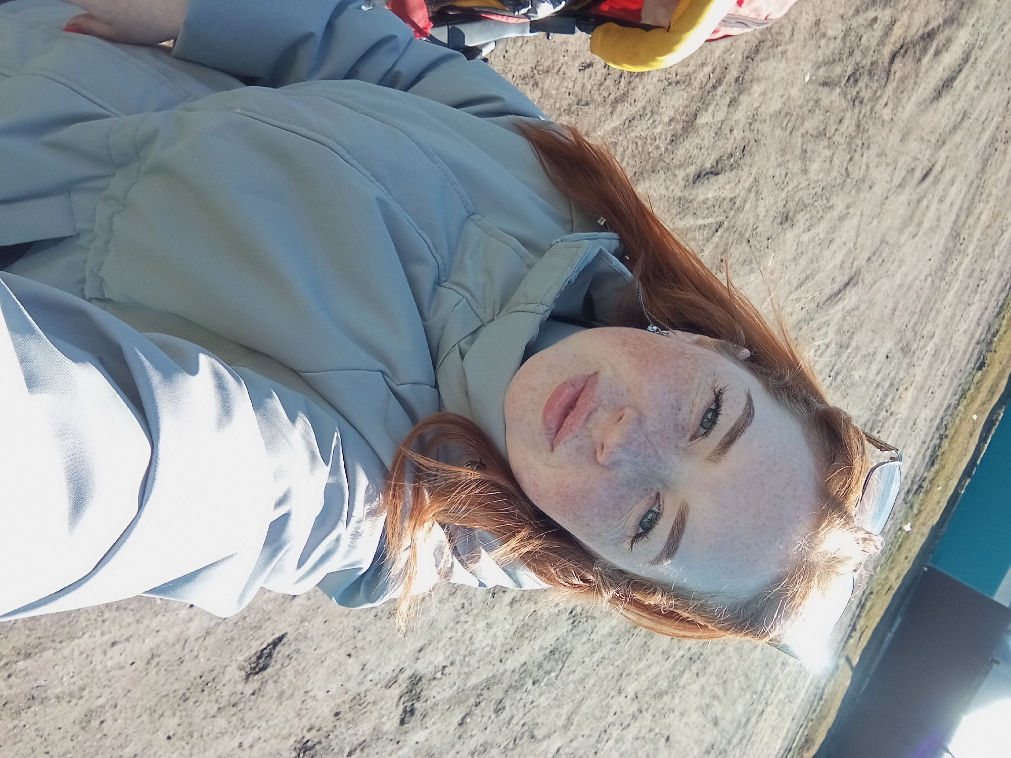 The image depicts a fair-skinned woman with red hair and freckles lying on a surface that appears to be sandy or dirt. She is wearing a light blue coat. A chain necklace is visible. Sunglasses are near her head. The location appears to be outdoors. The image was taken in Yurgamysh, Russia.