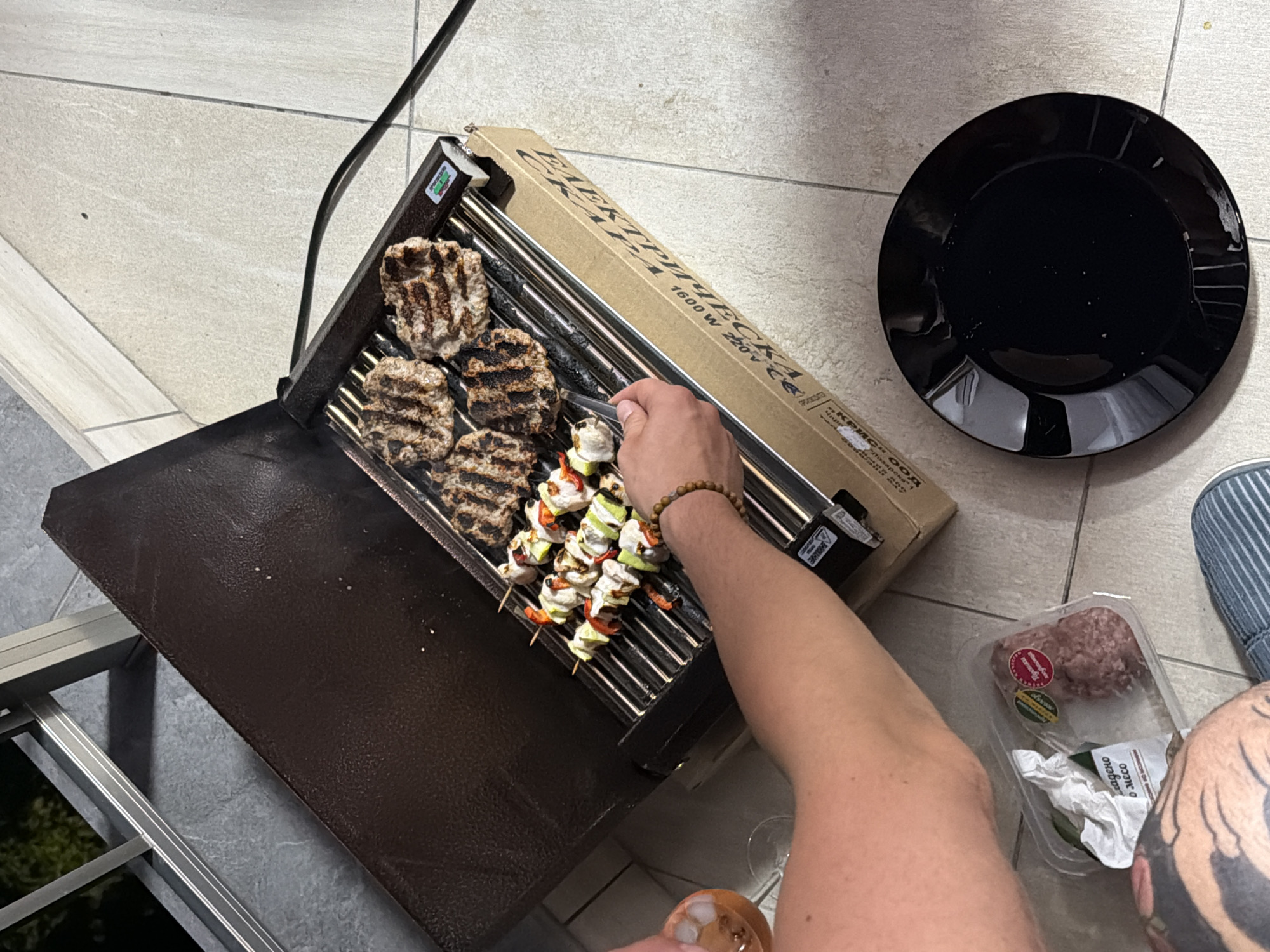 A person is grilling meat and skewers on a portable electric grill on a tiled balcony in Plovdiv, Bulgaria. The person is wearing a blue striped shoe and has a tattoo on their left leg. They are holding a fork and turning skewers with pieces of meat, vegetables, and possibly cheese. There are several grilled patties on the grill as well. A black plate sits on the floor next to the grill, and a container of raw ground meat is visible in the foreground. The background shows a balcony railing and foliage. The lighting suggests it is evening or late afternoon.