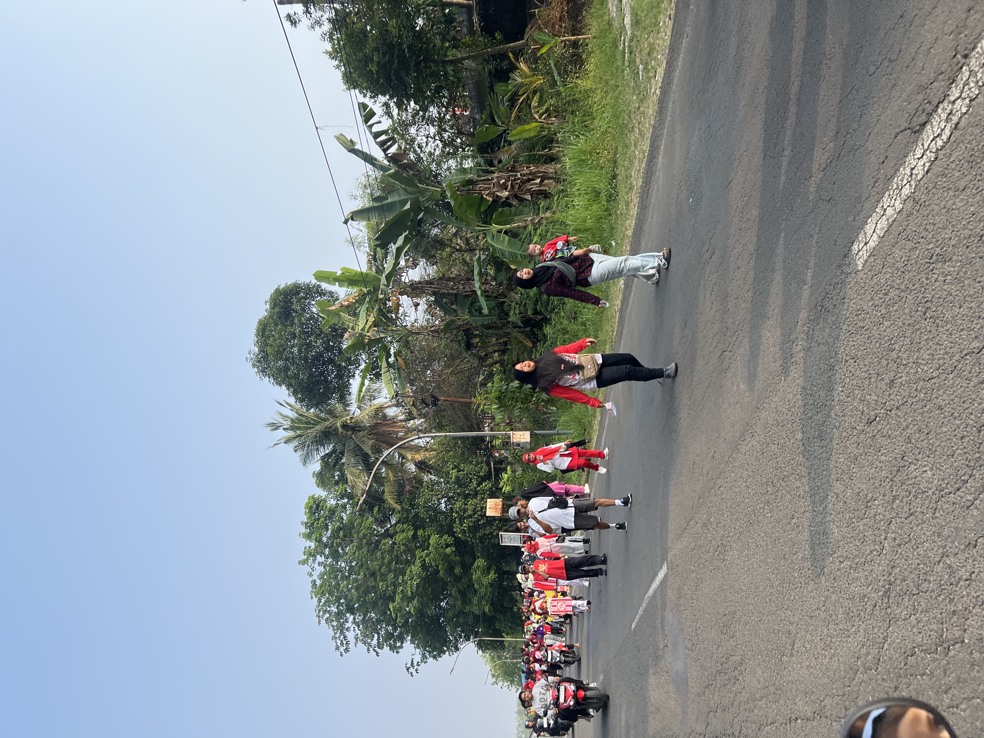 The image depicts a street scene in Cibinong, Indonesia, where a group of people are participating in a procession or walk. The road is paved and shows signs of wear, with a white dashed line marking the lane. On the right side of the road, lush green vegetation and banana trees are visible, suggesting a tropical environment. The sky is clear and blue, indicating fair weather.
A significant number of people are walking along the road, most of whom are dressed in red and white attire, common colors for national celebrations or sporting events in Indonesia. Some individuals are wearing headscarves. A few motorcycles are also present, with riders participating in the event. The people appear to be in motion, walking forward.
In the foreground, a motorcycle's side mirror is partially visible, reflecting a person's face. This suggests that the photograph was taken from the perspective of someone on a motorcycle. The overall atmosphere appears to be lively and communal, possibly a parade or a health walk.