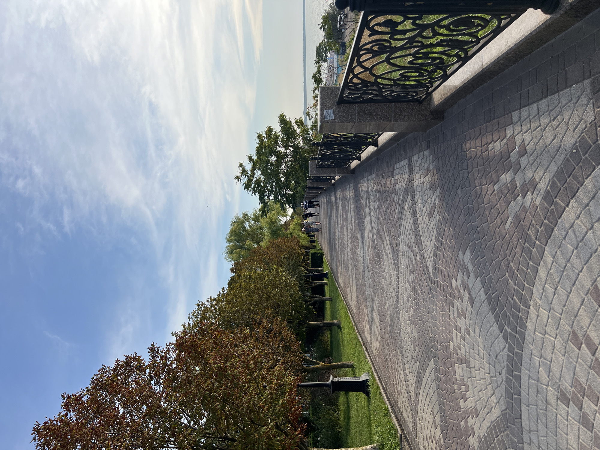 A paved walkway lined with trees and an ornate metal railing runs alongside a body of water. Several people are walking on the path, and the sky is blue with scattered clouds. The location is the Yeysk embankment in Yeysk, Russia.