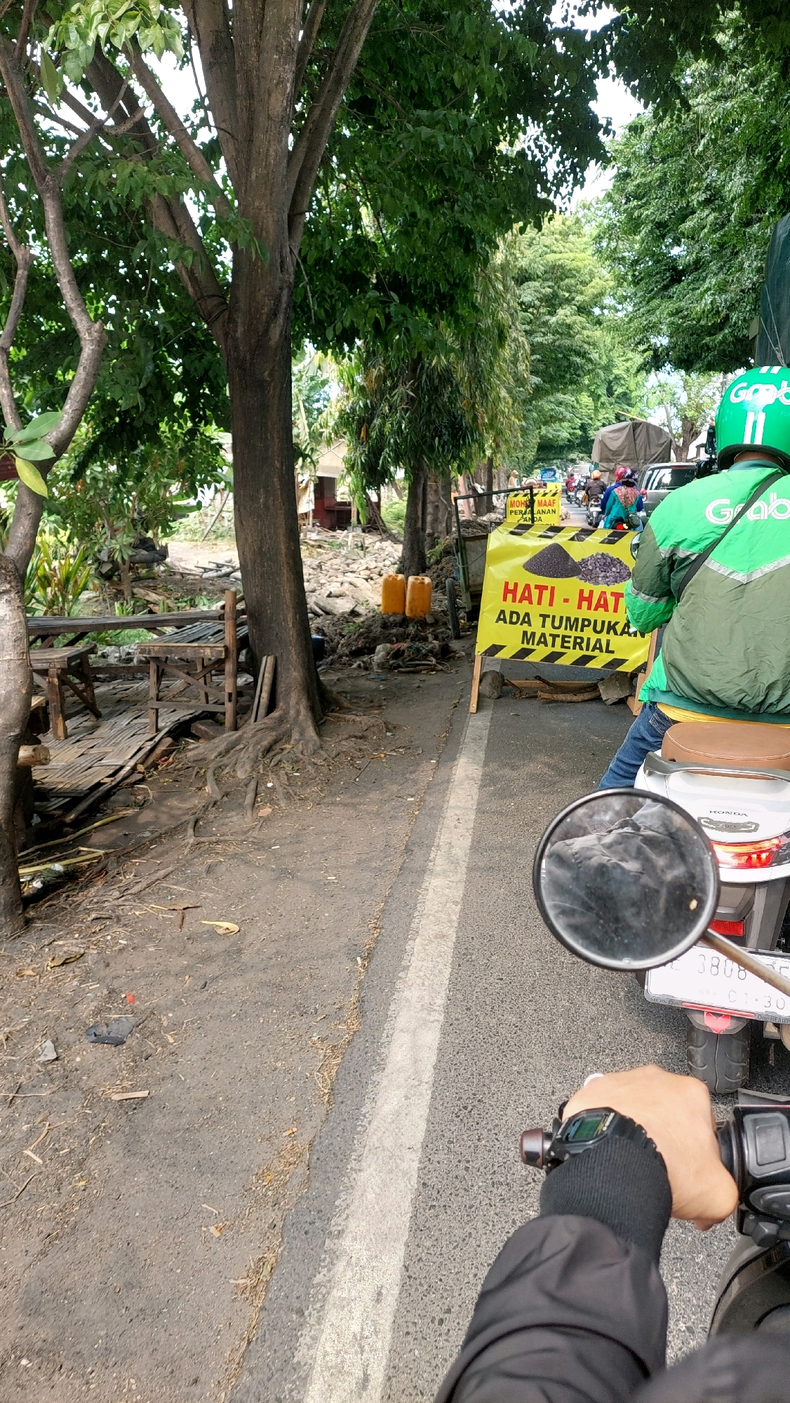 A first-person perspective captures a motorcycle rider's view on a road in Pilang, Indonesia. The rider's gloved hand grips the handlebars, with a black jacket sleeve visible. Ahead, a yellow sign with black and yellow stripes reads "HATI - HAT ADA TUMPUKAN MATERIAL," warning of a material pile. Behind the sign, two orange barrels and piles of gravel are visible. The road is asphalt with a white painted line. To the left, trees with dense green foliage line the road, and some wooden structures, possibly benches, are present. In the background, other vehicles, including motorcycles and a truck, are on the road, suggesting traffic. The overall environment appears to be an outdoor scene with vegetation and signs of construction or road work.