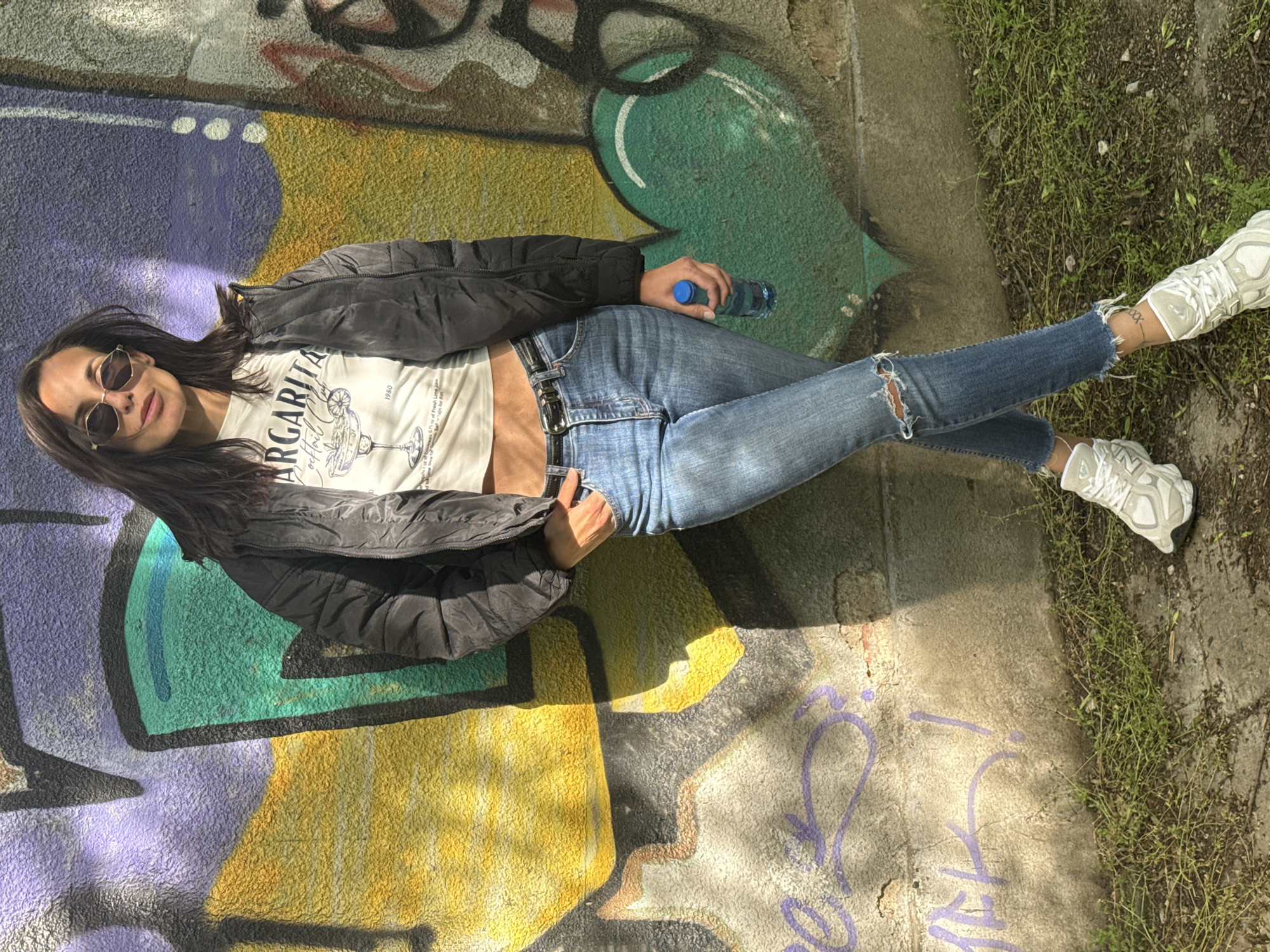 A woman is reclining on a graffiti-covered surface in an outdoor environment. She wears sunglasses, a white t-shirt with a "Margarita" graphic, a black jacket, blue jeans with a rip at the knee, and white sneakers. She holds a blue water bottle in her left hand. The ground is partly concrete with some patches of grass. The graffiti is multi-colored, with prominent purple, green, and yellow sections. The specific location within Pleven, Bulgaria, cannot be determined from the image.