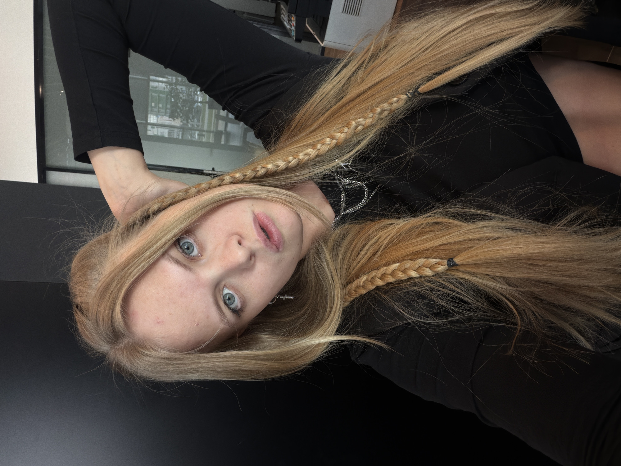 The image features a young woman with long, straight blonde hair and prominent light blue eyes, looking directly into the camera. Her head is tilted, giving the impression of an inverted perspective, as she appears to be lying down. Her fair skin shows a few subtle blemishes, and her lips are slightly parted.
Her long blonde hair is a dominant element, flowing around her head. Several thin, three-strand braids are woven into her hair on both sides, secured with small black elastic bands. She is dressed in a simple, long-sleeved black top or dress. Around her neck, she wears a silver-toned chain necklace featuring a stylized pendant that resembles an initial or a distinct symbol. Small silver earrings are also visible.
The background is predominantly dark, suggesting she is resting on a dark surface or against a dark backdrop. To the upper left of the image (her right), a window reflects an indistinct view, hinting at an indoor or outdoor environment with faint greenery and structural elements. A section of a white wall is also visible. To the upper right (her left), there are hints of dark furniture or equipment, possibly an air vent or part of a computer setup. The lighting is clear and soft, indicating an indoor setting, likely during daylight hours. No explicit text is clearly legible, apart from the stylized necklace pendant. This scene is located in Izhevsk, Russia.