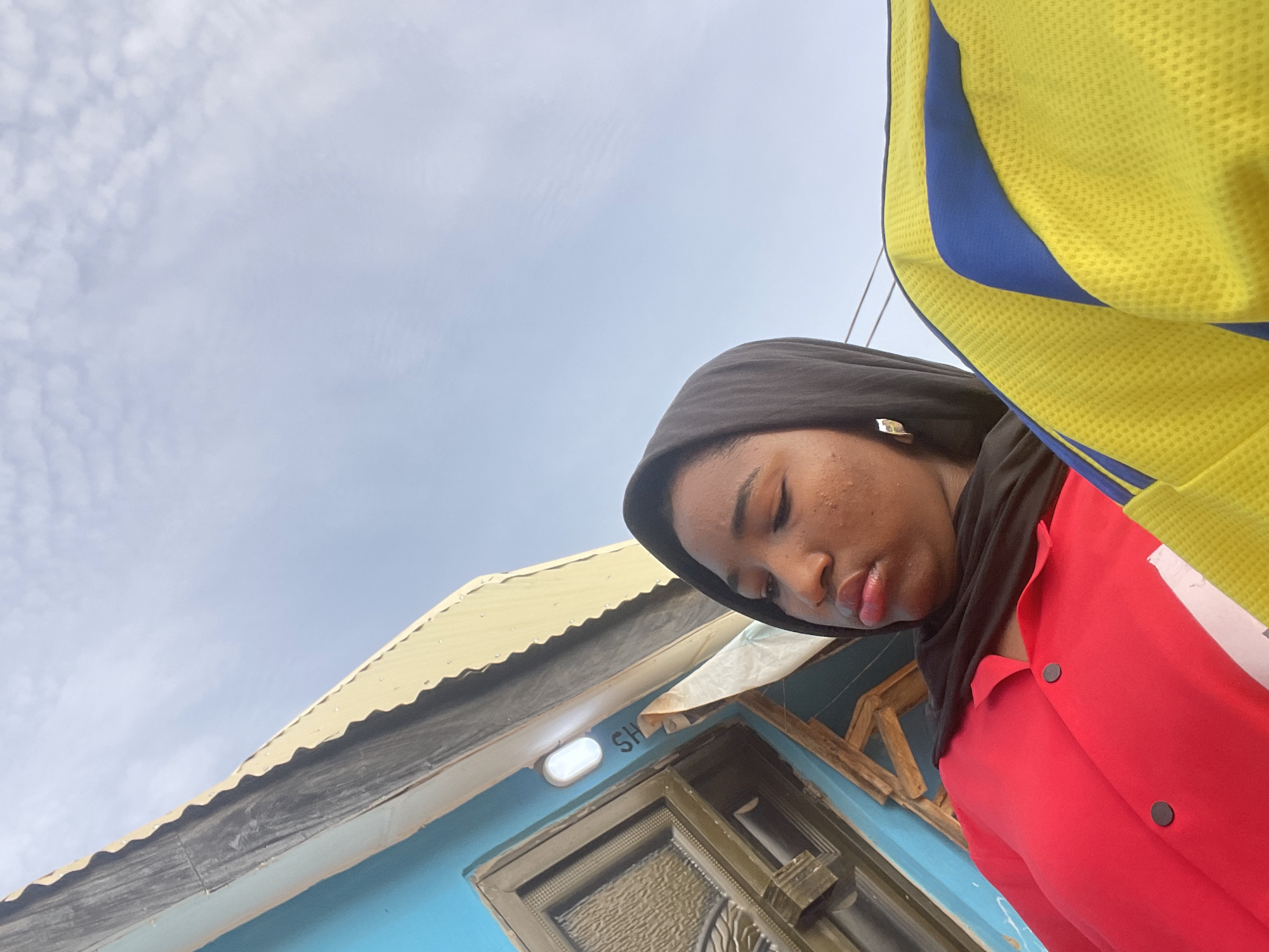 A low-angle, outdoor shot captures a person looking downwards, set against a bright, cloudy sky and a building in Lokoja, Nigeria. The person, positioned on the right side of the frame, is wearing a black head covering (hijab) and a vibrant red top or jacket with dark buttons. A section of a yellow garment with blue stripes or patterns is also visible at the top right, suggesting an additional layer of clothing or an accessory.
The person's eyes appear closed or heavily downcast, lending a serene or contemplative expression. Their lips are slightly parted, and some blemishes are visible on their cheek and forehead.
In the background, the upper portion of a building with a light blue wall and a corrugated yellowish-beige metal roof is prominent on the left. A dark, possibly metal, door with intricate patterns is visible on the building's facade. Above the door, next to a white rectangular light fixture, the partial letters "SH" are discernible on the blue wall. The sky, occupying the upper and central parts of the image, is bright and filled with scattered white clouds, indicating daytime. The perspective is taken from below, looking up towards the subject and the sky.
