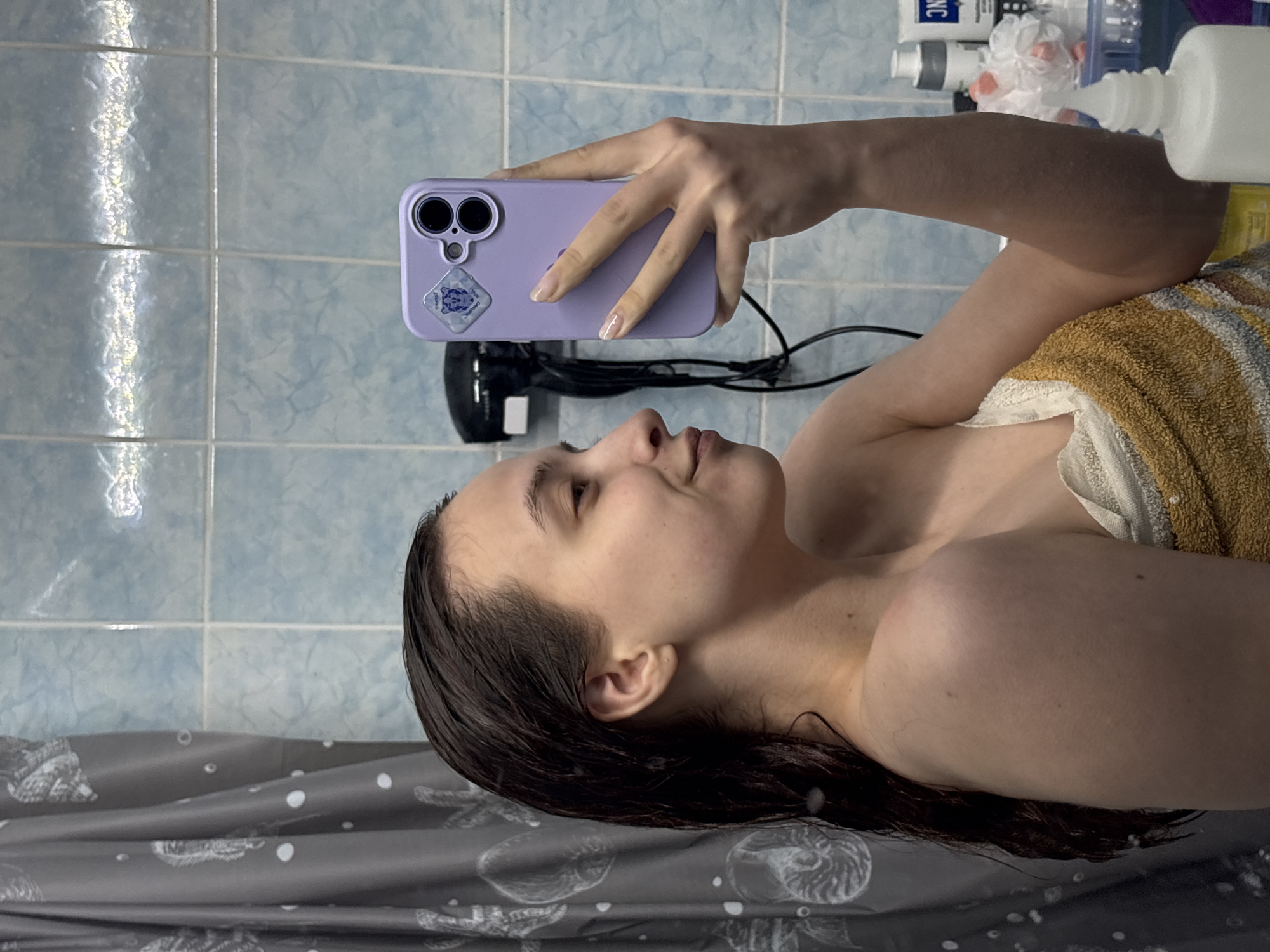 A woman with wet, dark brown hair is taking a selfie in a bathroom with blue tiled walls. She is holding a purple phone in her right hand, which is slightly extended. The phone has a sticker on the back and a dual-camera setup. She is wrapped in a white and gold striped towel. Behind the phone, a black hairdryer with a white nozzle is visible. Above the dryer are various toiletries on a shelf. The woman is standing in a shower with a grey and white patterned curtain. The location might be Прикамский, Russia, given the presence of Russian text on some of the toiletries in the background.