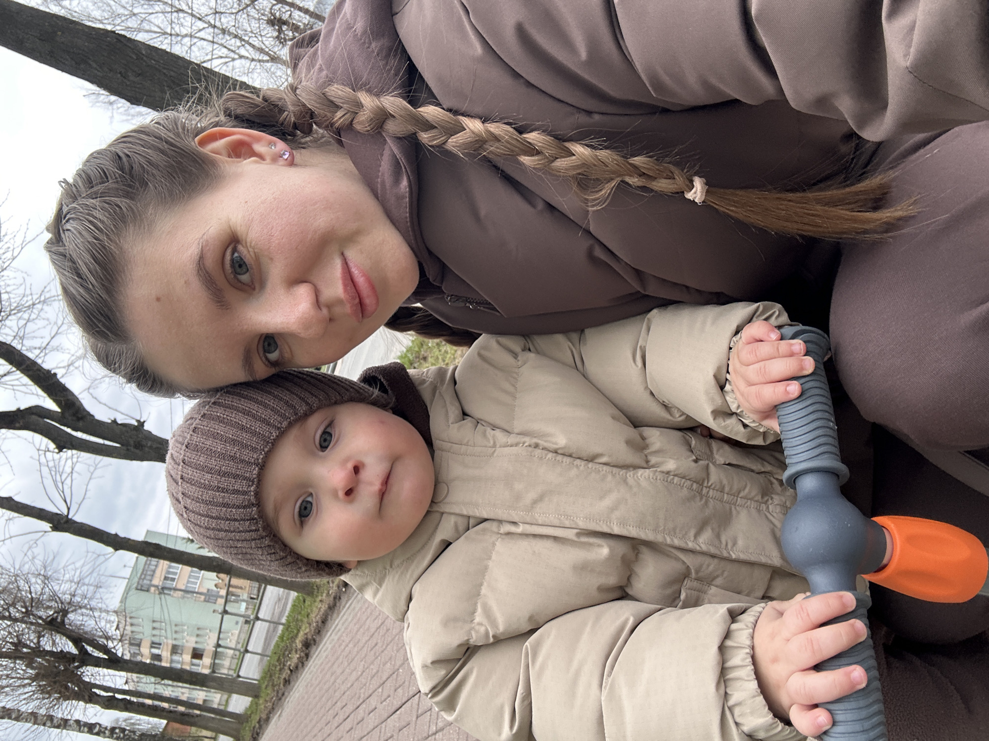 A woman and a toddler are posing for a selfie outdoors. The woman has light skin, a braid, and is wearing a brown puffy jacket. The toddler, wearing a brown knit hat and light beige jacket, holds a gray and orange scooter handle. They appear to be in a park with bare trees and a building in the background. The setting is determined to be Klin, Russia based on contextual clues, which may include architectural styles and landscape features. The weather seems overcast.