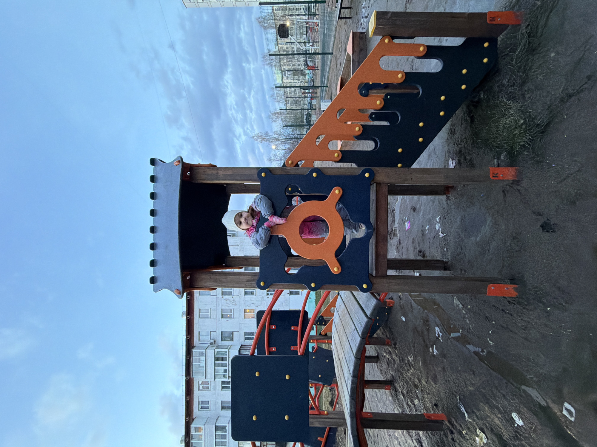 A young girl is pictured sitting on a colorful playground structure in an outdoor urban setting. She is positioned in an opening that features an orange steering wheel-like design, looking directly at the camera with a neutral to slight smile. The girl wears a white knit hat, a pink scarf, a gray jacket, and pink pants/tights, suggesting cold weather.
The playground equipment is made of wood and plastic components, primarily dark blue/black and orange, with yellow circular accents. It includes a castle-like top, steps, and a slide.
In the background, a large, light-colored apartment building with numerous windows and balconies is visible. Beyond a fence, bare trees and what appears to be a sports court or another section of the residential area can be seen. The ground around the playground is dark, muddy, and wet, indicating damp conditions, possibly from recent rain or melting snow. The sky is overcast and light blue with scattered clouds, suggesting a dull, cloudy day, likely in late autumn, winter, or early spring. The overall scene conveys a typical day in a residential area of Chaikovsky, Russia. There is no clearly visible text.