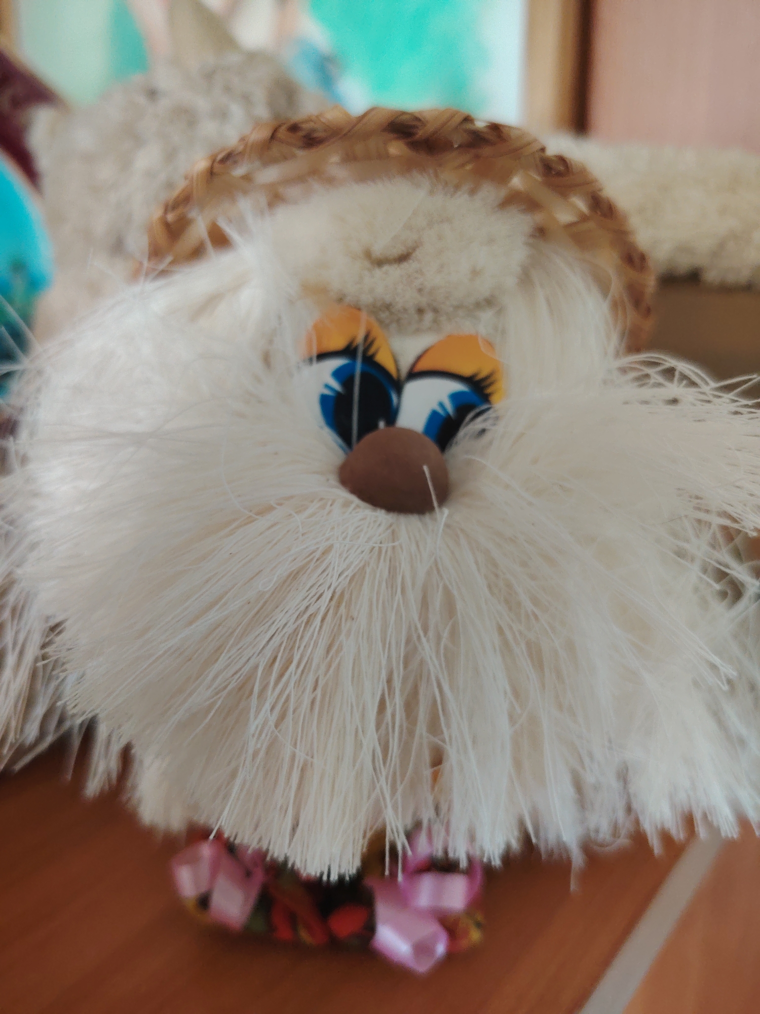 The image displays a close-up, vertically oriented, eye-level photograph of a handcrafted doll. The doll is predominantly white and furry, with large, cartoonish eyes featuring blue irises and orange rims. A small, brown, ovular object serves as its nose. A woven straw-like band encircles the doll's head, resembling a halo or headdress. The lower portion of the doll is adorned with pink ribbons and what appear to be decorative beads or charms. The doll is positioned on a wooden surface, which is likely a table or shelf. The background is out of focus, showing muted colors and shapes that suggest an indoor environment, possibly a home. There are no people visible in the image, and no specific landmarks or identifying features place this scene within Konakovo, Russia.