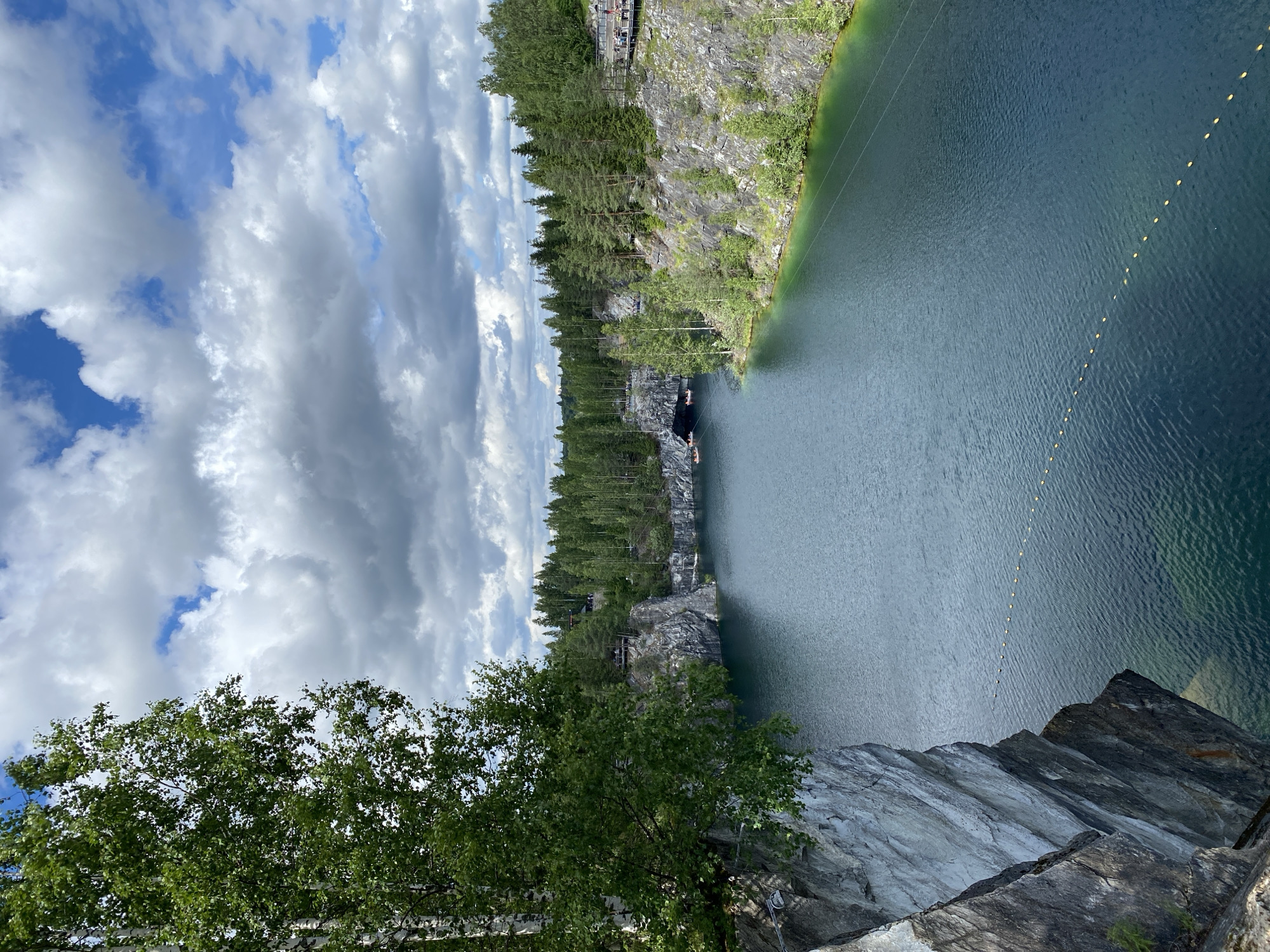 The image depicts the Marble Canyon, a flooded former quarry located in Ruskeala, Russia. The canyon features steep, sheer marble cliffs plunging into deep, clear turquoise water. Dense evergreen forests line the upper rim of the canyon, with some deciduous trees, including birches with their white bark, visible on the left foreground. The sky is partly cloudy with dramatic grey and white cumulus clouds interspersed with patches of blue.
In the water, several small boats are visible, some appearing to be navigating towards or within a cave opening in the far left cliff face. A line of yellow buoys stretches across the water, suggesting a designated swimming or boating area. On the right side of the canyon, a wooden boardwalk is visible, elevated along the cliff, with people walking and observing the scenery. The overall impression is of a natural landscape transformed by quarrying, now a popular tourist destination. The lighting suggests it is daytime, likely late morning or afternoon, under a dynamic sky.