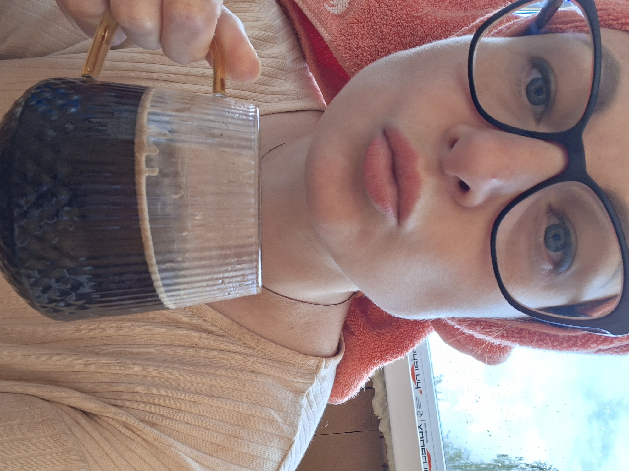 A woman wearing a pink towel on her head and black-rimmed glasses is holding a glass mug filled with a dark liquid, possibly coffee. She is making a puckered expression with her lips. The background appears to be a window with rain streaks and a blurry outdoor scene. The woman is wearing a light-colored, ribbed shirt. The setting is likely an indoor environment, possibly a home, in Забалоцкі сельскі Савет, Belarus.