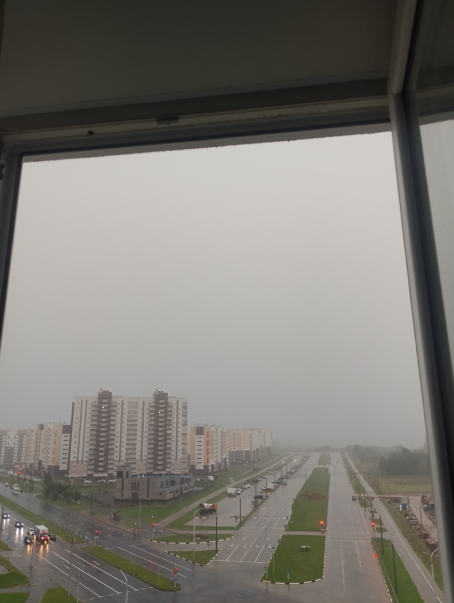 This is a high-angle, outdoor, full shot of a modern city street scene on a foggy day, viewed through a window frame. The foreground is dominated by the window frame and a light gray sky. In the mid-ground, a wide, wet asphalt road with visible lane markings cuts through the scene. Traffic lights are visible, showing red. Several vehicles, including cars and trucks, are present on the road, some with their headlights on, indicating movement. The road is flanked by green grassy medians and sidewalks. On either side of the road, there are numerous multi-story apartment buildings, mostly light-colored with a modern architectural style. Some buildings have visible windows and balconies. In the distance, the fog obscures the rest of the urban landscape and any natural elements beyond the buildings. The overall impression is one of a suburban or developing urban area under overcast and misty weather conditions.