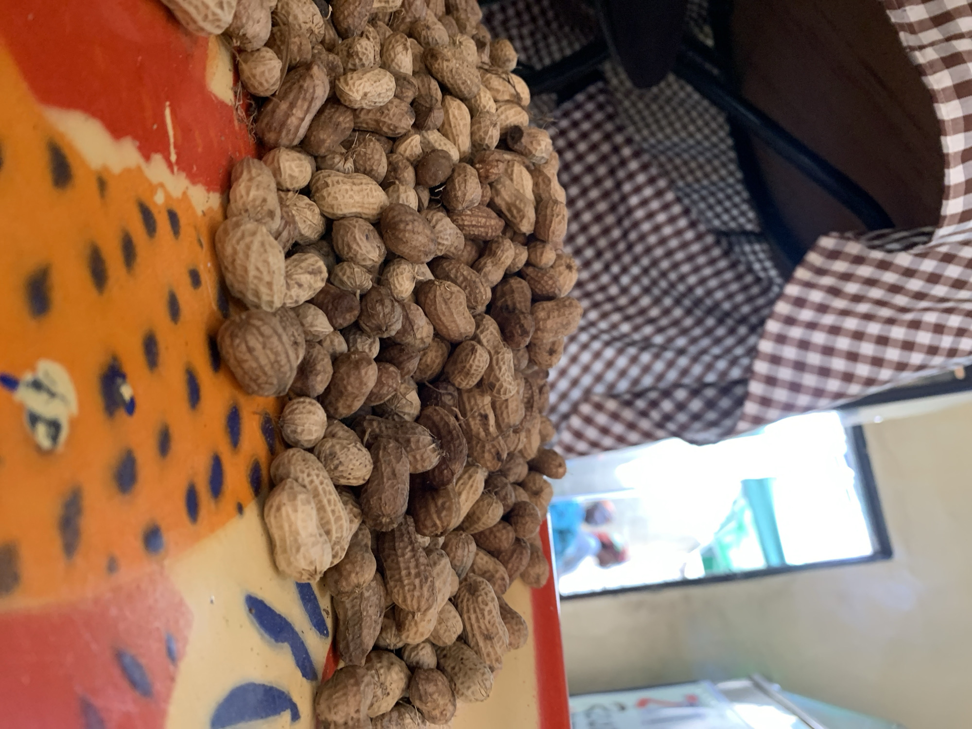 A low-angle, close-up photograph depicts a substantial mound of unshelled peanuts positioned on a multicolored, patterned surface. The peanuts display varying tones of light and dark brown, with discernible shell textures. The underlying surface is an abstract design featuring segments of orange, red, and off-white, embellished with irregular dark blue markings. In the upper left background, a brown and white checkered fabric is visible, positioned below a dark brown surface, likely the underside of a table, with black metal support structures present. The central and right background features a brightly illuminated opening, through which two blurry human figures are discernible; one figure appears to be wearing blue, and the other has a red or orange head covering. To the far right, a vertical object, potentially a sign, with faint red and white details is partially visible. This scene is located in the City of New Stadium, Nigeria.