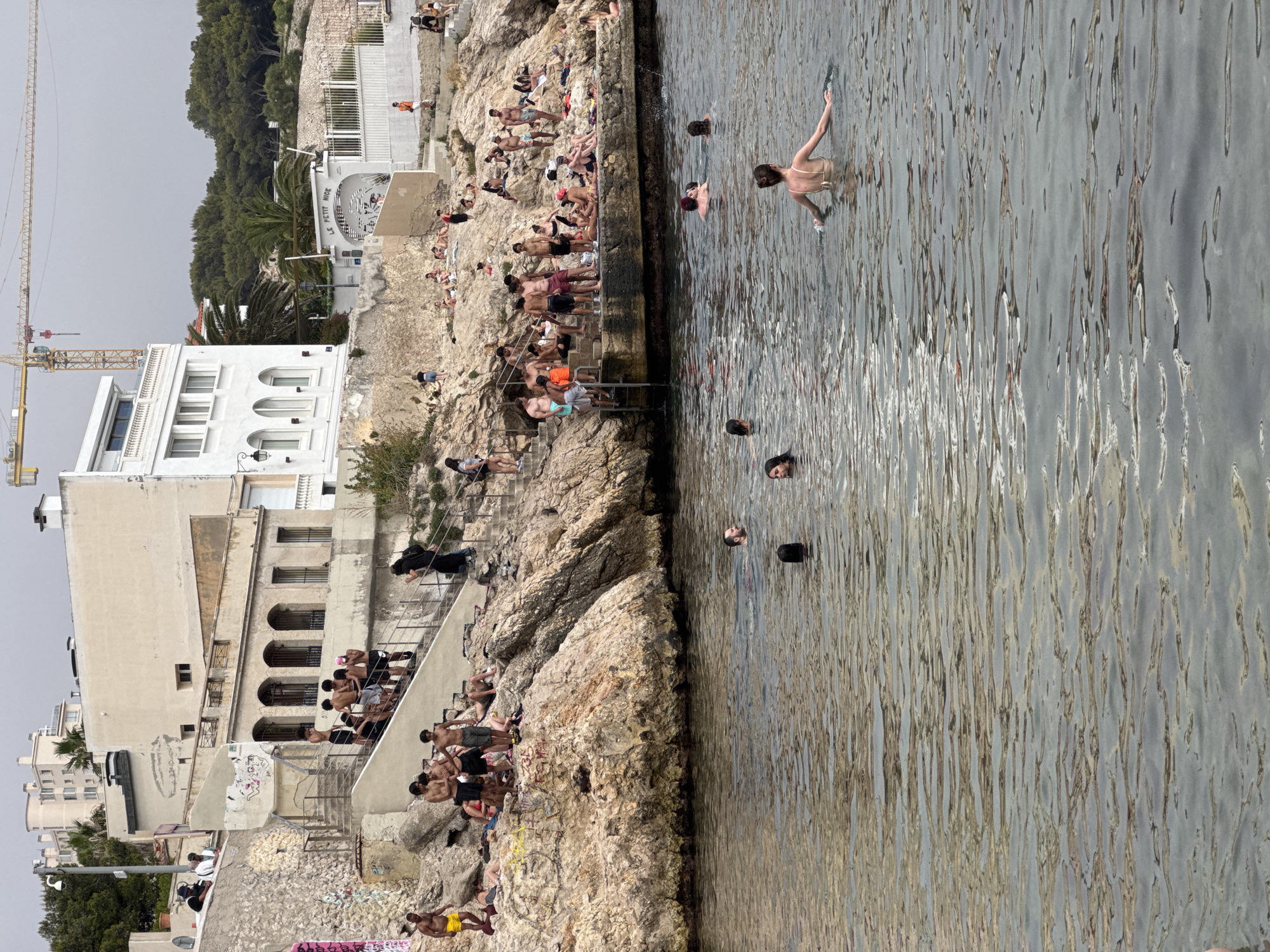 The image shows a crowded outdoor swimming area carved into rocky terrain, possibly a natural cove or a man-made pool by the sea. The scene is vibrant with activity on a bright, likely sunny day, judging by the strong shadows and the numerous people enjoying the water and sun.
The subjects are primarily people of various ages, many in swimwear, engaging in swimming, sunbathing, and socializing. Some are in the water, while others are seated or standing on the rocky ledges and stairs that lead down to the water. A few individuals are walking up and down the stairs, carrying towels or bags. The water itself appears clear and reflects the sunlight, creating a shimmering surface.
Notable details include the architecture of the buildings visible in the background. One building, with an arched entrance, is labeled "LE PETIT NICE." Another larger, pale-colored building with multiple windows and arched openings is also prominent. The presence of a construction crane in the upper left corner suggests ongoing development or infrastructure work in the vicinity. The overall atmosphere appears lively and relaxed, with people enjoying a summer day at the coast.
The terrain is rocky and uneven, with some areas covered in sparse vegetation. Graffiti is visible on some of the stone walls and structures, adding a touch of urban edginess to the natural setting. The time of day appears to be midday or early afternoon, given the angle of the shadows. The location cues, specifically the architecture and the name "LE PETIT NICE," strongly suggest this is a popular spot in Marseille, France.