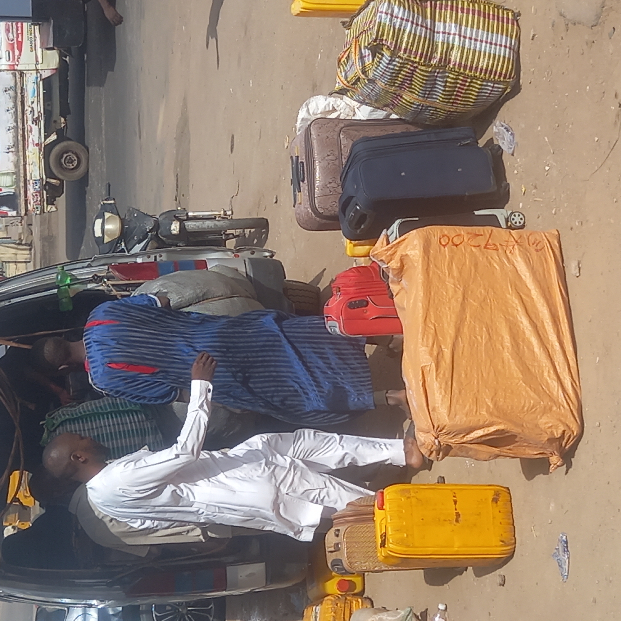 This media file captures an outdoor scene in Dadi Riba, Nigeria, likely a roadside or transportation hub, depicting activity related to travel or transport. The setting appears bright and sunny, indicating daytime.
In the foreground and center, two men are lying down amidst various goods and luggage. One man wears a long, loose-fitting blue garment with red stripes and is positioned near the back of what appears to be a vehicle, possibly a truck or large car. Another man, dressed in a white long-sleeved shirt and white trousers, lies further down, partially obscured by items. Their relaxed postures suggest resting, waiting, or being transported.
The scene is dominated by a substantial amount of luggage and cargo. Prominently visible are several suitcases: a brown patterned one, a black one with wheels, and a red one with wheels. A large, checkered woven bag with yellow, red, and white stripes is also present. Many items are covered by tarps, including a large orange tarp on which the handwritten text "¥7200" is visible in red. Other items include several grey or white sacks, yellow jerry cans, and a green plastic bottle.
A white vehicle, possibly a pickup truck or commercial van, is partially visible on the left, with one of its wheels and some side markings. A black motorbike is parked nearby. The ground is dusty or sandy. The overall impression is one of a busy, yet momentarily still, travel or cargo loading/unloading point.