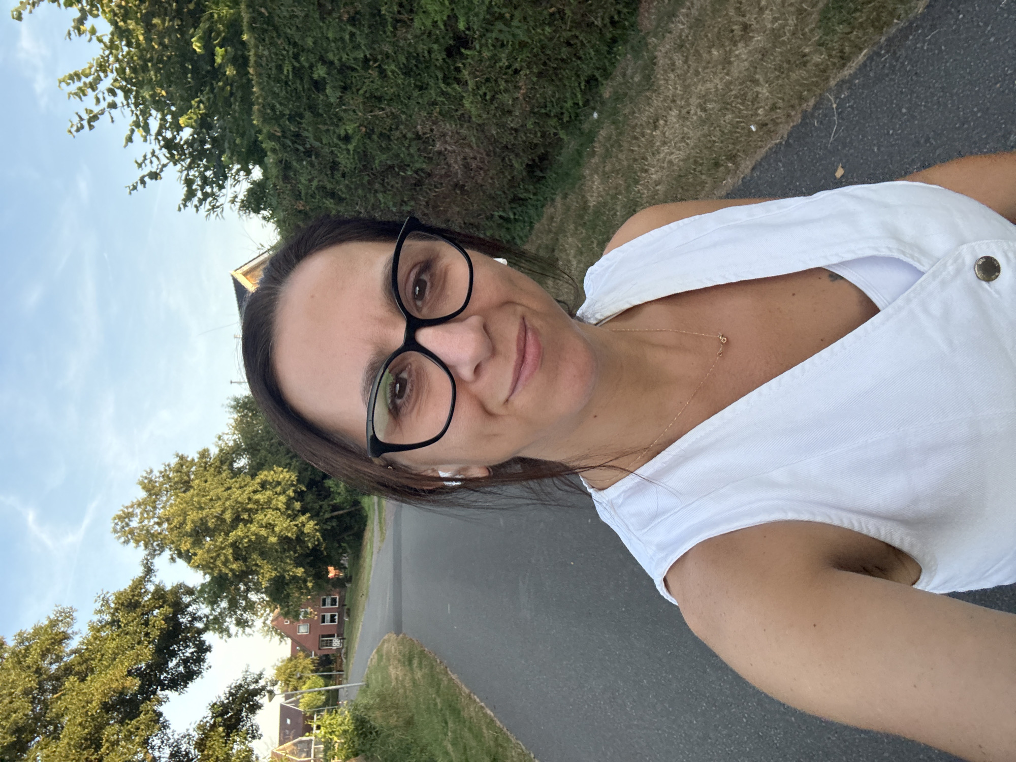 In Marum, Netherlands, a woman is captured in a selfie on a paved path. She is wearing a white sleeveless top and black-rimmed glasses. In her ears are white wireless earbuds. A thin gold necklace with a small charm is visible around her neck. The path curves to the left and is bordered by green grass and vegetation. In the background, there are trees, houses, and a green hedge. The sky above is blue with scattered white clouds.
