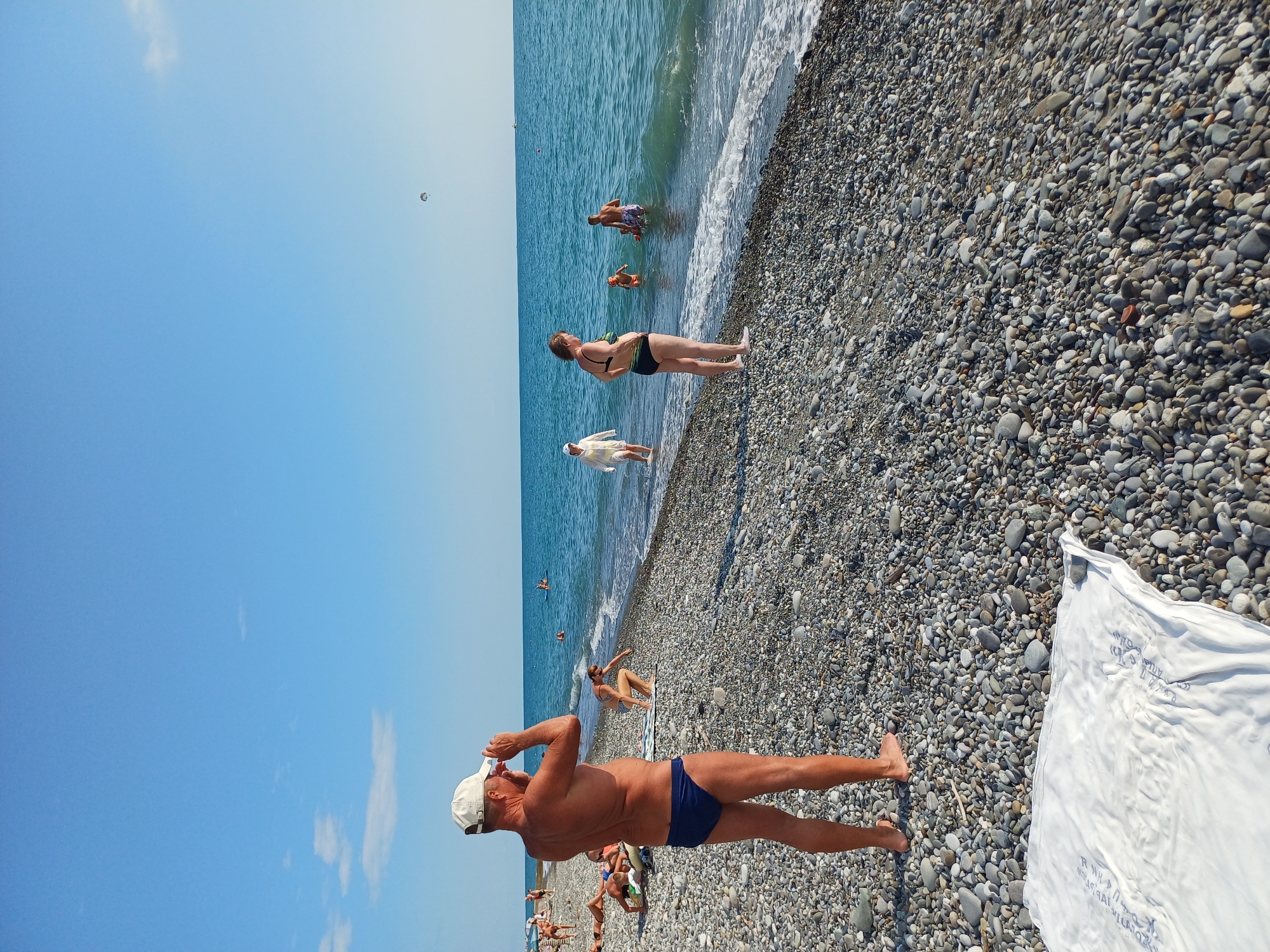 The image depicts a sunny day at a pebble beach in Sochi, Russia. Several people are visible, engaged in various beach activities. In the foreground, a man in blue swim trunks and a white cap is adjusting his cap. Further down the beach, a woman in a bikini is sitting on the pebbles, and in the water, several people are swimming or wading. The sea is blue and appears calm with gentle waves. The sky is clear and blue, with a few wispy clouds. The ground is covered in smooth, grey pebbles. A white fabric, possibly a beach towel or sheet, lies on the pebbles in the lower right corner of the image. The overall mood is relaxed and summery, indicative of a typical day at the beach. No visible text is present in the image.