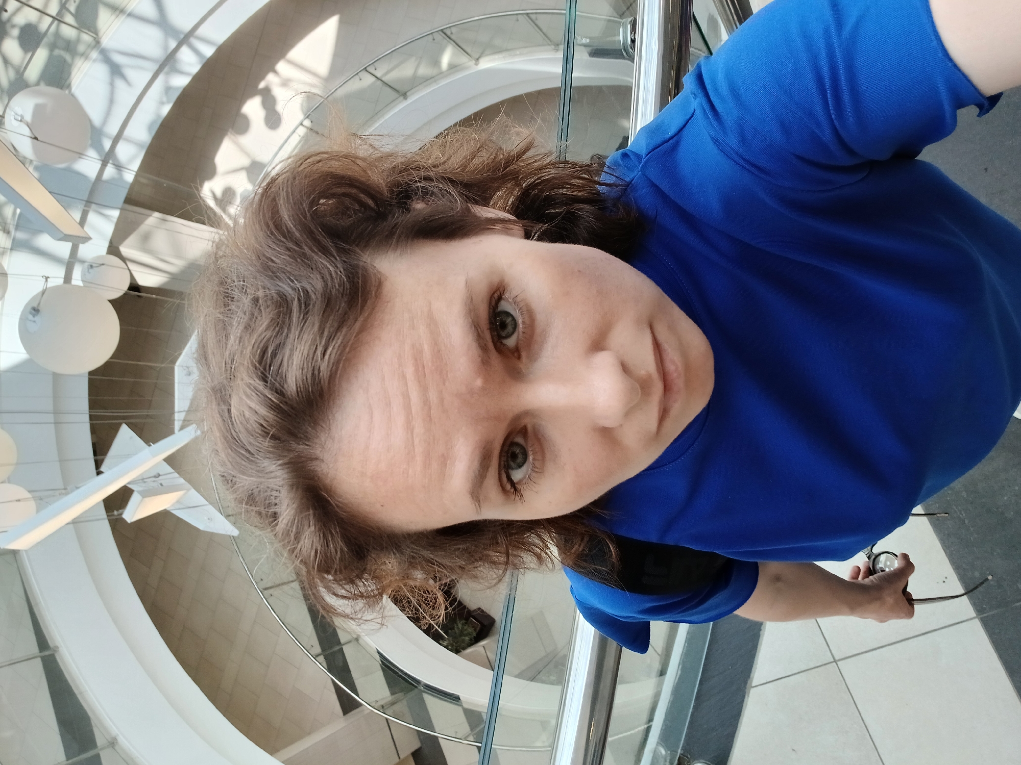 A woman is taking a selfie from a high vantage point, looking directly into the camera. She has wavy brown hair and is wearing a bright blue t-shirt. Her left hand is visible holding a pair of eyeglasses. The background appears to be an interior atrium or shopping mall with a multi-level, curved architectural design featuring white structures and glass railings. The lighting suggests a bright, daytime setting. The environment appears to be a modern, indoor public space.