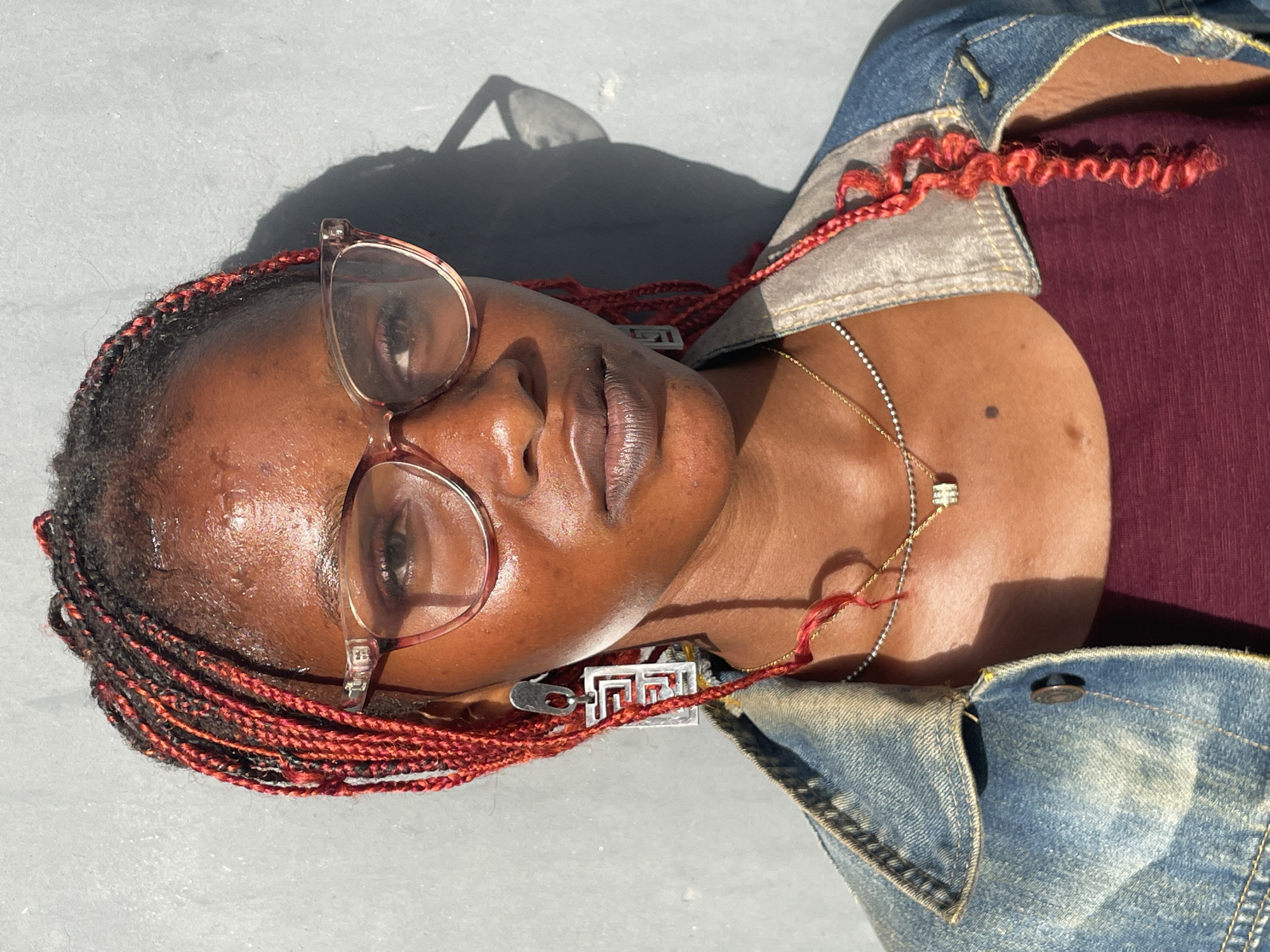 The image presents a close-up, overhead view of a person with dark skin, lying horizontally on a light gray, textured surface that appears to be concrete. The person wears glasses with translucent, light-colored frames and has numerous red braids, some with black intertwining, framing their face. Their eyes are closed or looking downwards, and their lips are slightly parted.
They are dressed in a blue denim jacket with visible stitching over a dark maroon or deep red top. Two necklaces adorn their neck: a delicate gold chain and a silver-toned chain featuring a small, square, sparkling pendant. A unique silver-toned pendant, characterized by an intricate geometric, maze-like design, is clipped or woven into the red braids near the left ear.
Bright sunlight creates sharp shadows of the person's head and glasses on the surface behind them, suggesting clear, sunny weather, likely around midday or early afternoon. The skin on the forehead and lips appears somewhat glistening, indicating possible warmth or humidity. The overall expression conveyed is one of calm or contemplation. There is no discernible text visible in the image. The setting is an outdoor location, consistent with Badagry, Nigeria.