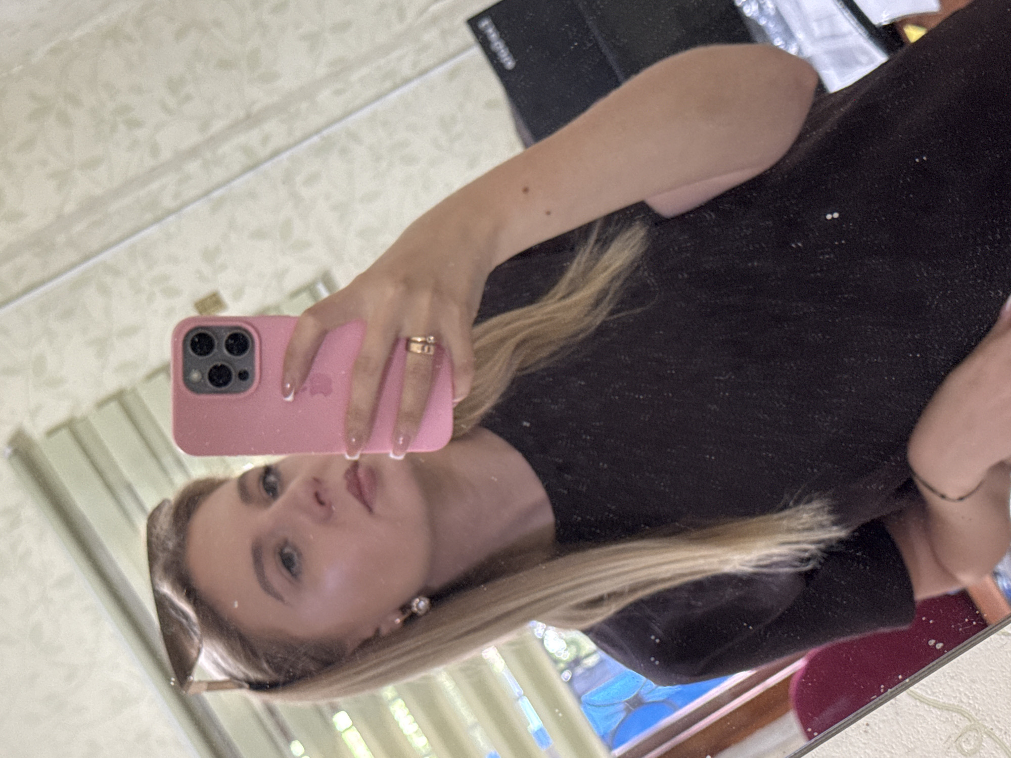 A woman is taking a mirror selfie in a room. She is wearing a black t-shirt, sunglasses on top of her head, and jewelry. She is holding a pink smartphone in her right hand, which is pointed towards the mirror to capture the reflection. Her hair is blonde and straight. The background shows a patterned wallpaper and blinds.