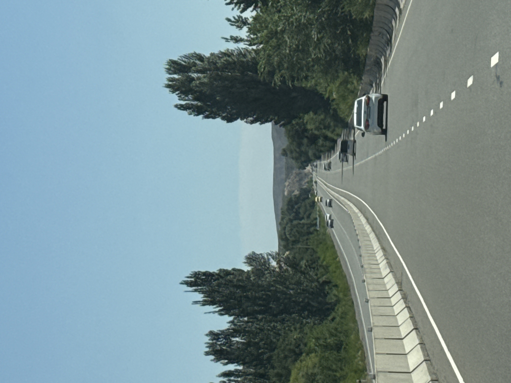 A vertical, eye-level, medium shot captures a highway stretching into the distance under a clear blue sky. The road is paved and features a white line marking the lanes. On either side of the highway, tall, dark green trees stand in dense formations, creating a natural border. Some of these trees are unusually slender and pointed, resembling cypress trees. The landscape beyond the trees appears to be hilly or mountainous, with a muted color palette of greens and browns. Several vehicles are visible on the highway, all moving in the same direction. In the foreground, a white car is prominent, followed by other vehicles further down the road. Concrete barriers line the left side of the highway. The scene appears to be set in a rural or semi-rural area. The location is identified as Unknown, Kyrgyzstan.