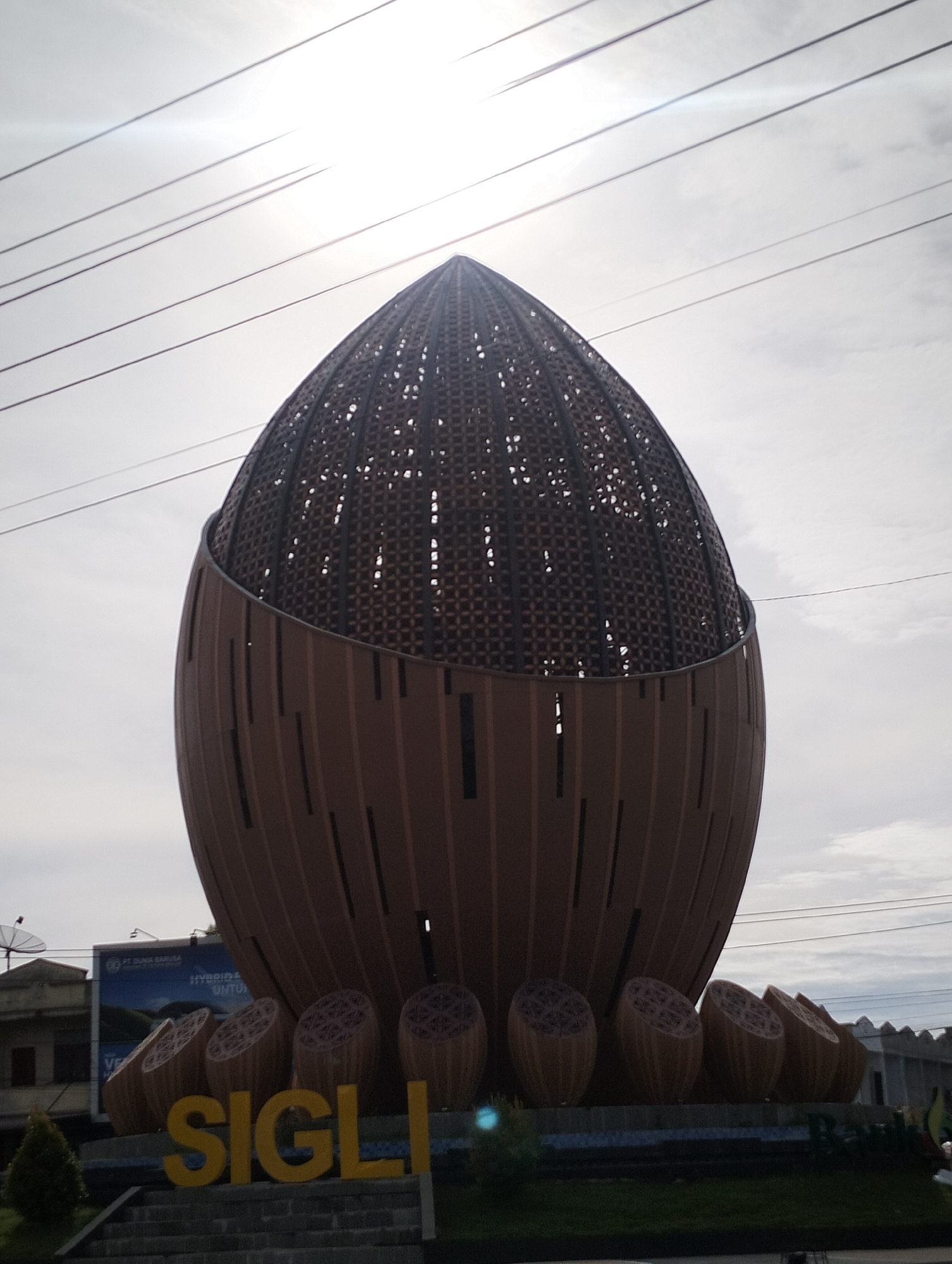 The image depicts a large, oval-shaped monument or structure in Lampeudeu Baroh, Indonesia. The monument is primarily brown and features a latticework design at the top and vertical slats on the lower portion. The base consists of multiple smaller, similarly designed oval shapes. The word "SIGLI" is spelled out in yellow letters in front of the monument. Overhead power lines crisscross the sky, which is overcast. In the background, there are buildings and a billboard with text and an image. A small amount of greenery is visible at the base of the monument.