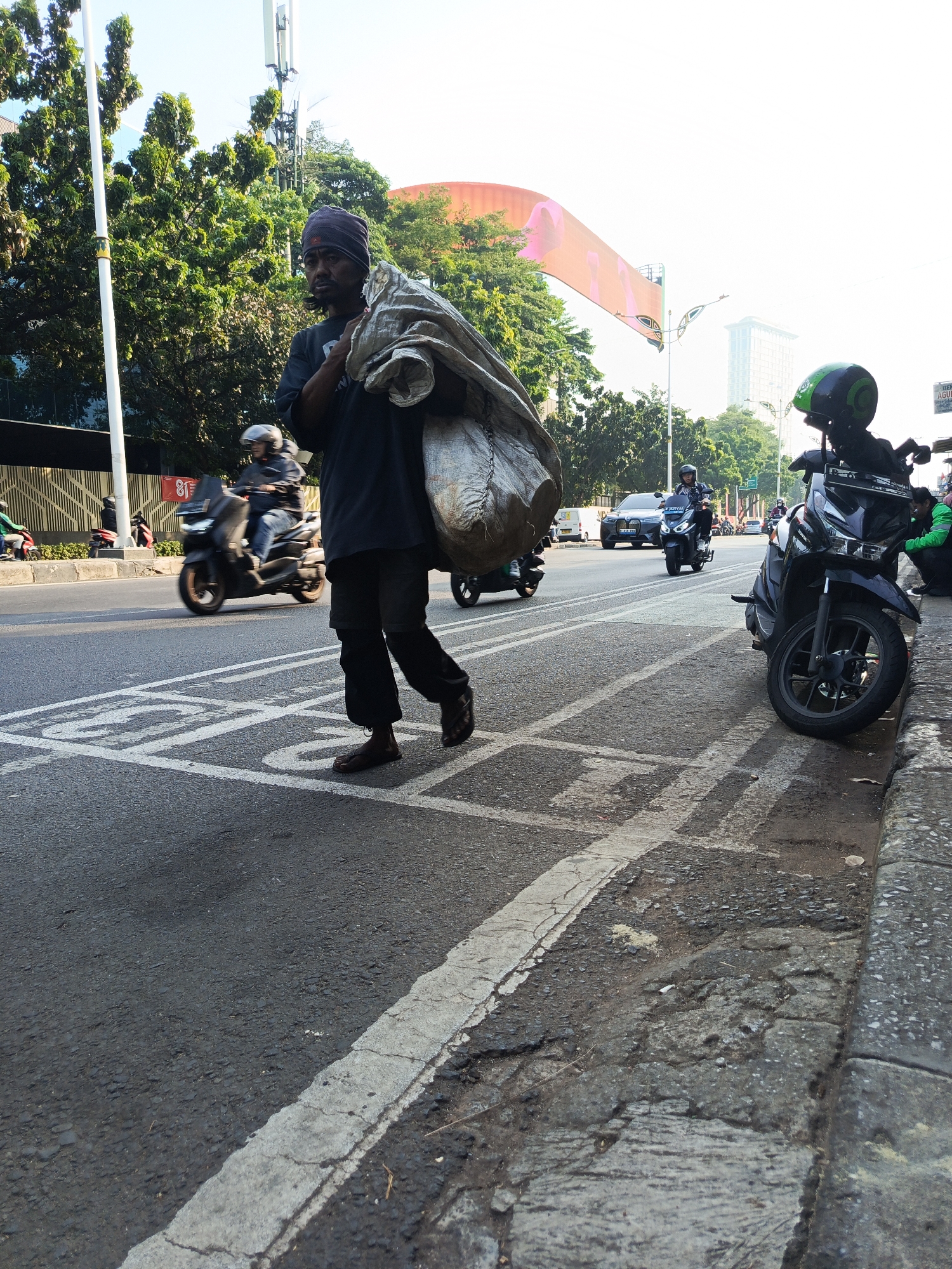 A man walks along a city street in Central Jakarta, Indonesia, carrying a large sack of material over his shoulder. He is wearing dark clothing and a head covering. The street is paved and appears to be busy with traffic, including motorcycles and cars. Trees and buildings line the street, and a large banner or advertisement is visible in the background. The scene is illuminated by natural daylight.