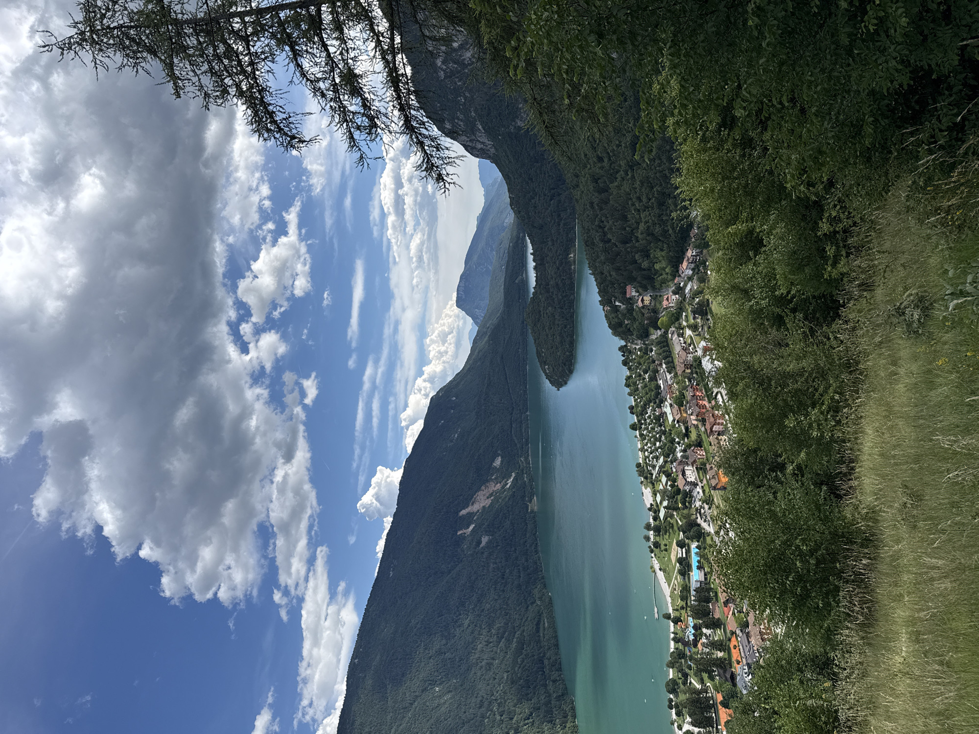 The image shows a high-angle view of Lake Molveno in Molveno, Italy. The lake is a vibrant turquoise color and is surrounded by steep, forested mountains. A small town with red-roofed buildings is nestled along the lakeshore, and a campground is visible with tents and RVs. The foreground is a grassy hillside with some trees. The sky is blue with scattered white clouds. There are no people visible in the image. The overall scene is one of natural beauty and a tranquil vacation destination.