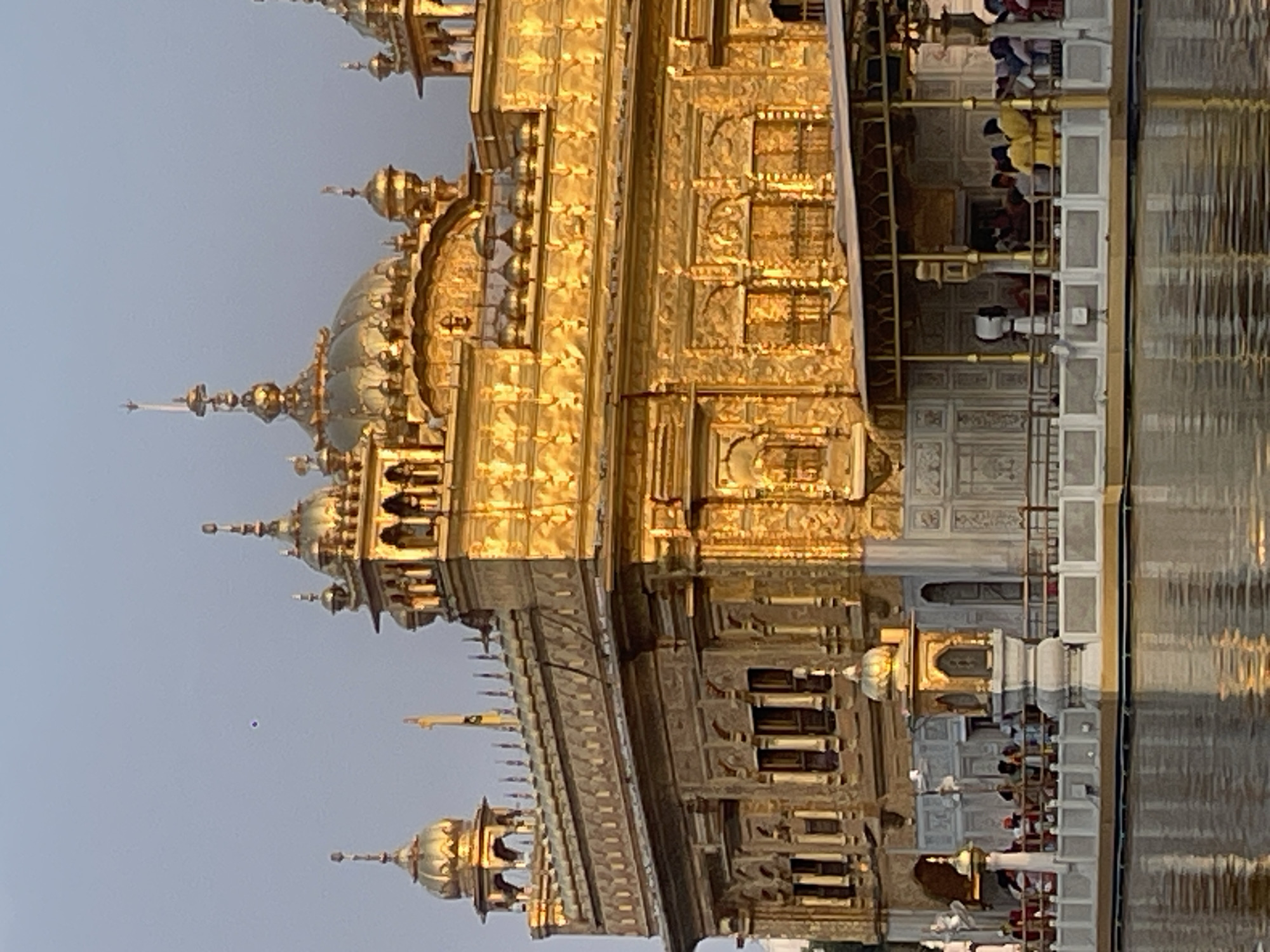 The image shows the Golden Temple in Amritsar, India. The temple is a Sikh Gurdwara and is one of the holiest sites in Sikhism. The temple is made of gold and is surrounded by a pool of water. In the image, people can be seen walking around the temple and on the bridge that leads to the temple. The sky is a light blue and the sun is setting, casting a warm glow on the temple. The temple is intricately decorated with carvings and ornamental work. The water in the pool reflects the golden structure, creating a stunning visual. The scene appears peaceful and serene.