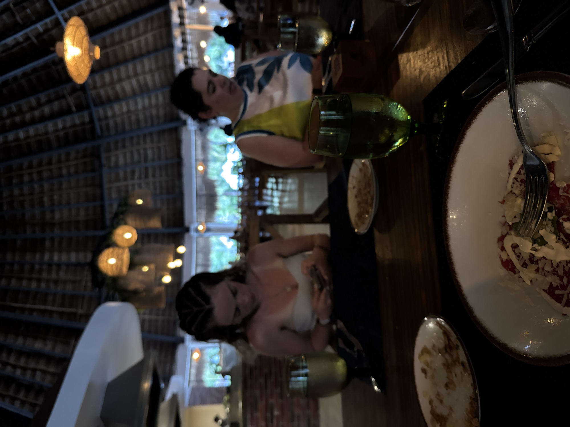 Two women sit at a wooden table in an indoor restaurant in Playa del Carmen, Mexico. The restaurant is under a thatched roof, with hanging lights. One woman wears a white and blue top with a yellow accent. The other woman is wearing a white top. A table setting includes plates, forks, and green-tinted glasses. A partially eaten salad is visible on one of the plates.