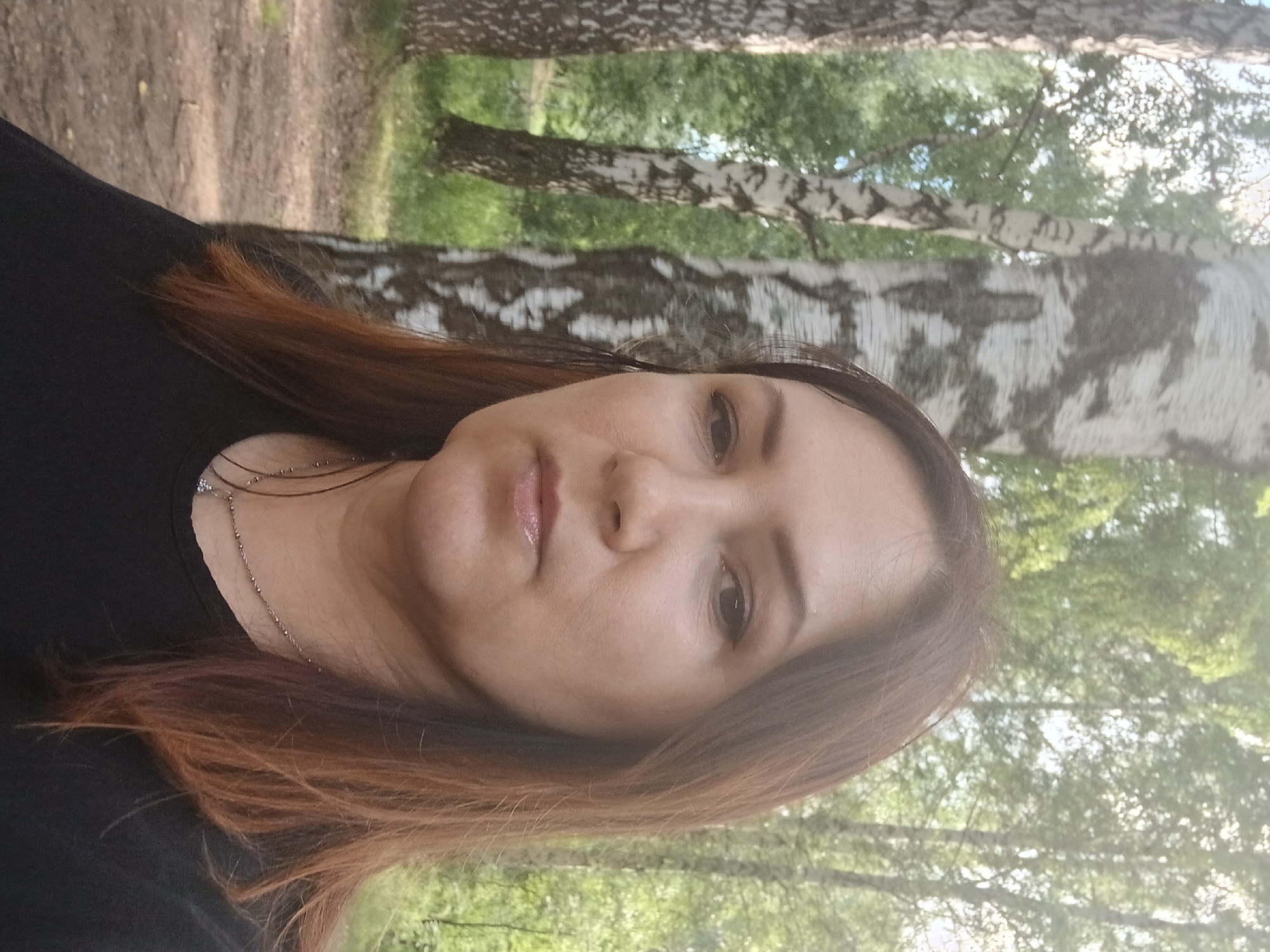 The media file is a portrait of a woman with auburn hair, taken outdoors. She is wearing a black top and a delicate silver necklace. Her expression is neutral. The background features birch trees with their characteristic white bark and green foliage, suggesting a park or forest setting. The lighting indicates it is daytime, likely late morning or afternoon, with dappled sunlight filtering through the leaves. There is no visible text in the image. The scene evokes a sense of tranquility and a connection with nature. The orientation of the image is sideways, with the woman's head on the right side of the frame.