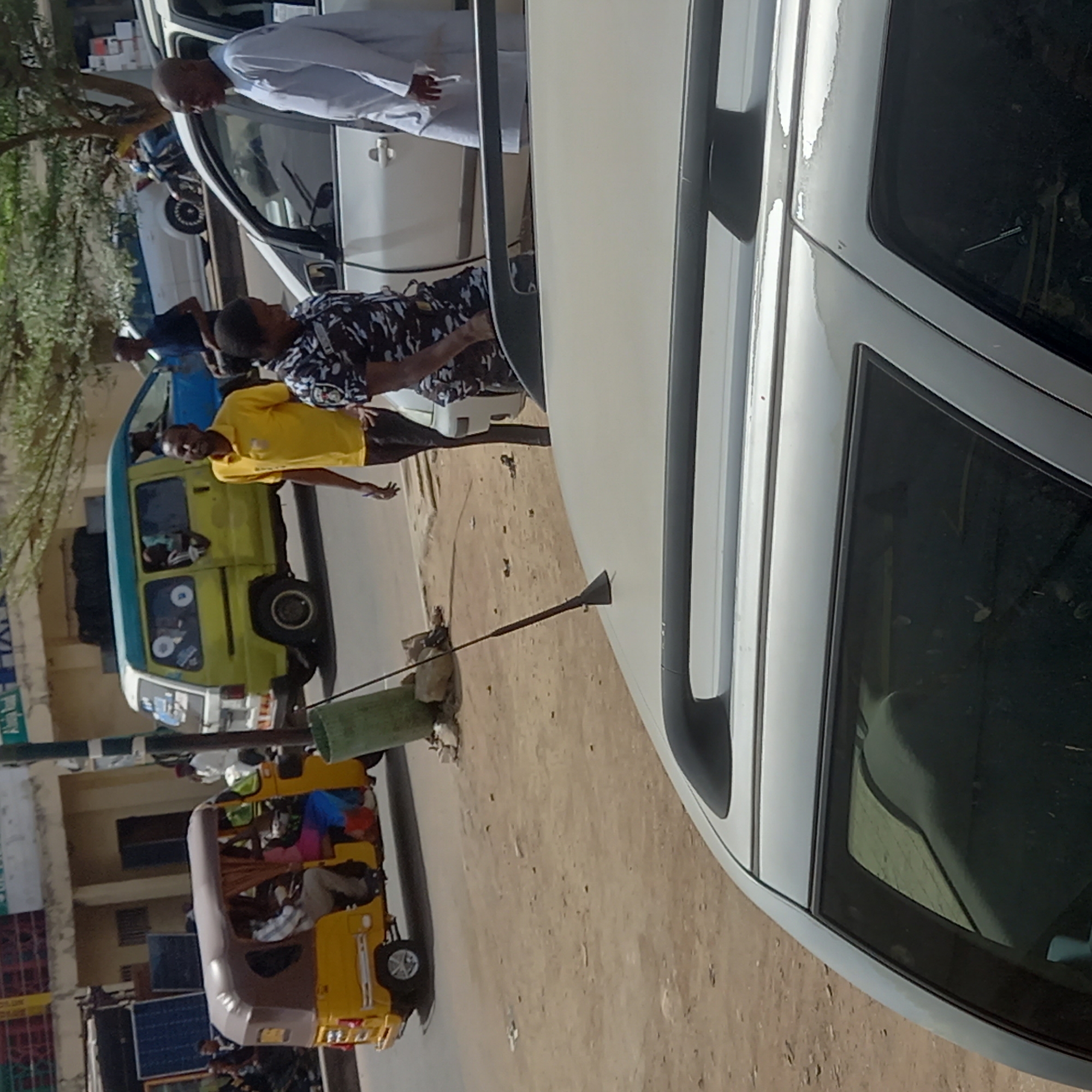 This image captures a busy street scene in Sardauna, Nigeria, under bright daylight. The scene is oriented vertically but depicts a horizontal street.
In the foreground, a white car, possibly a sedan or SUV, occupies the right side of the frame. A man in a white traditional robe (likely a thobe) is seen leaning into the driver's side window of this car. Beside him, another man in a black and white patterned shirt stands near the vehicle.
To the left of the white car, on a dirt or unpaved road, stands a man in a bright yellow t-shirt, looking towards the viewer. Behind him is a yellow and green mini-van or small bus, typical of public transport in the region. Further down the street, a yellow and brown auto-rickshaw, commonly known as a "keke Napep" in Nigeria, is visible, carrying several passengers.
Notable details include a long pole with a bell-shaped top, possibly an antenna or makeshift sign, stuck into the ground near a green bucket. Trees and foliage are visible on the far left. In the background, buildings line the street, and solar panels can be seen near one of the structures. The weather appears sunny and clear. No discernible emotions are evident from the individuals. Visible text on vehicles and buildings is indistinct and unreadable. The street itself appears well-trafficked, with various forms of local transportation present.