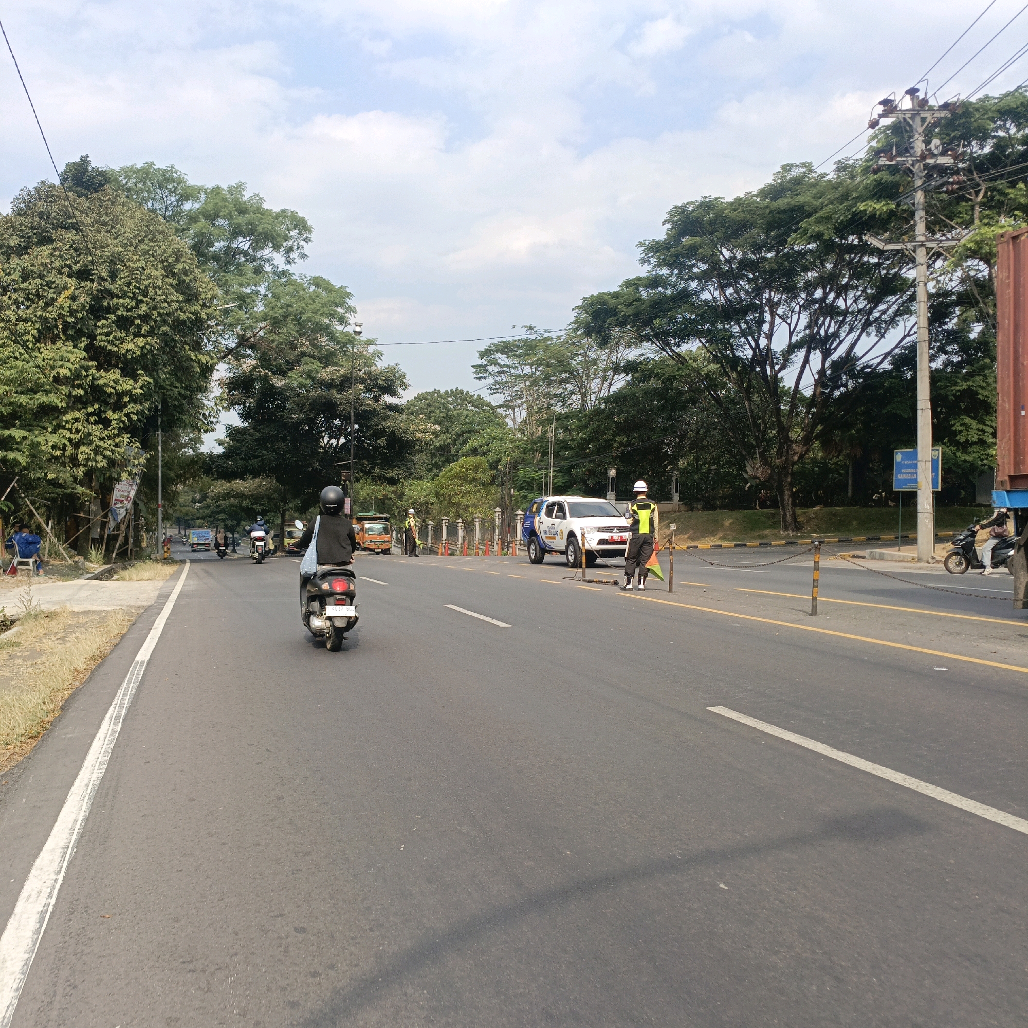 The image depicts a street scene in Salatiga, Indonesia. In the foreground, a person rides a scooter, viewed from behind. The scooter has a license plate that reads "4037 BBL". The road has a white line marking the edge of the lane and a yellow line further out. On the right side of the road, a white SUV with blue and yellow markings is parked, and two individuals in reflective vests and hard hats are standing near it, one holding a green flag. In the background, there are trees, traffic cones, and other vehicles including trucks and motorcycles. A utility pole with wires stands on the right side of the road. The sky is partly cloudy.