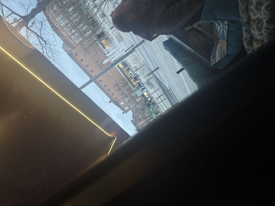 The image shows a blurry, angled view from inside a building, possibly a cafe or restaurant. A man's head is visible in the upper right, suggesting he's seated at a table. Outside, a city square is seen through a window, featuring buildings and what appear to be metallic structures, possibly public art. The lighting is dim inside, with a bright yellow streak from an unseen light source reflecting on the window. The overall image quality is poor, with a large portion of the foreground in dark shadow.