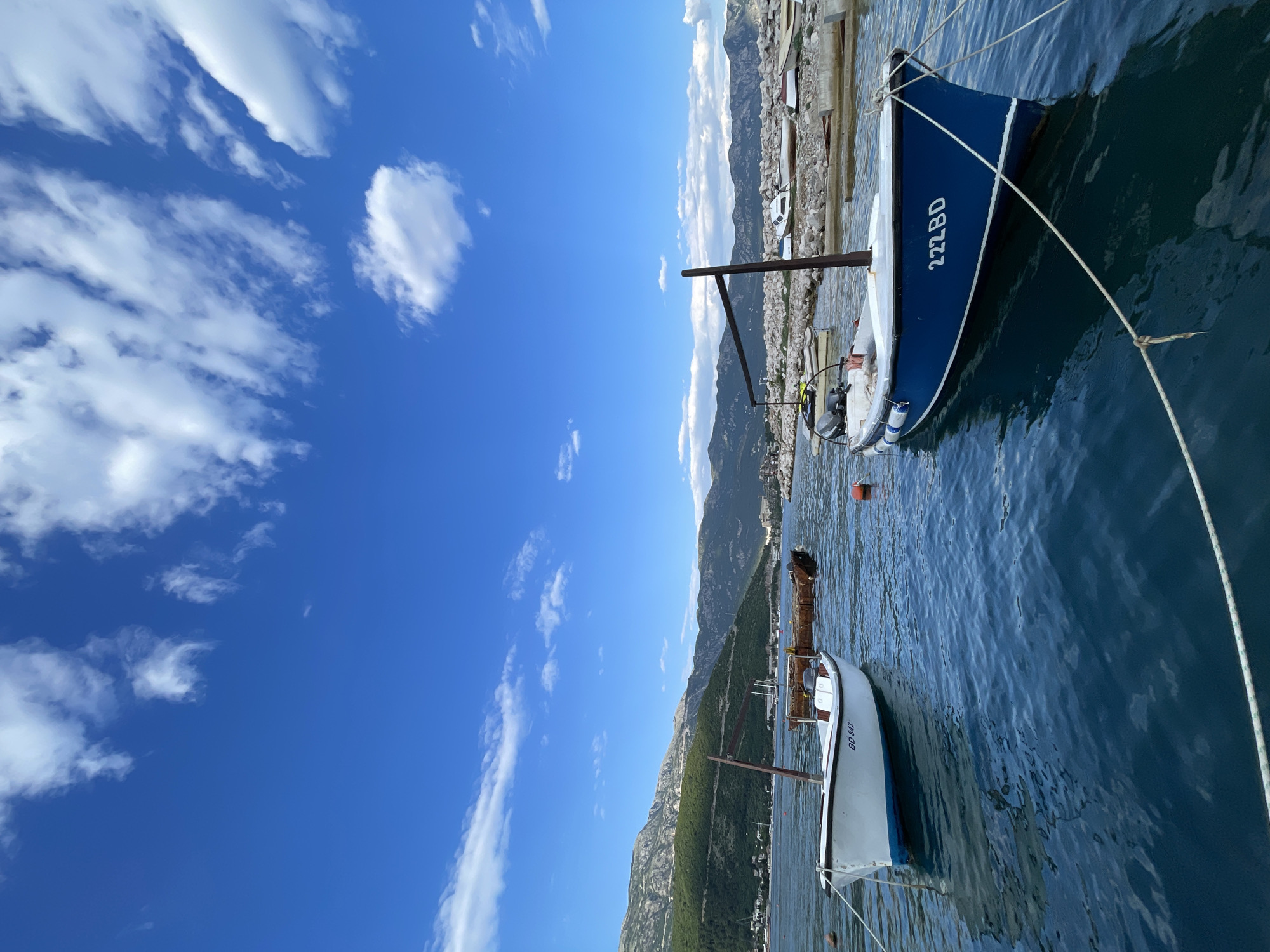The image shows a harbor scene with two boats in the foreground. The first boat, closer to the viewer, is white with blue accents, and it bears the inscription "BD 842". The second boat is a deeper blue with the inscription "222 BD". Both boats appear to be moored in the water.
In the background, there's a mountainous landscape with green vegetation covering the slopes. Buildings and structures are visible at the base of the mountains, indicating a settlement or town. The sky is a bright blue with scattered white clouds.