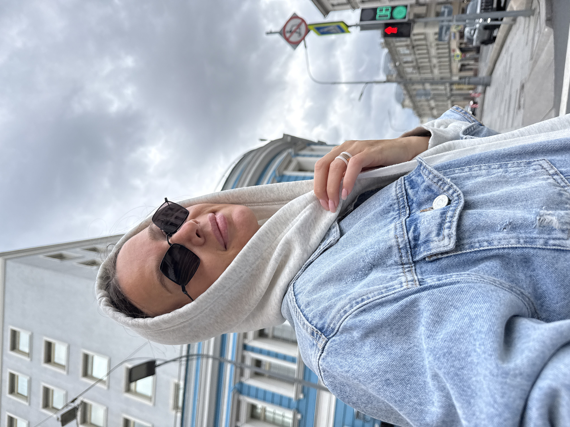 The image is a close-up, waist-up selfie of a woman in a light blue denim jacket and a grey hooded sweatshirt. She is wearing black, angular sunglasses and a ring on her left hand. Her hood is pulled up, framing her face. The background shows a cloudy sky and a city street with buildings and traffic lights. The architecture of the buildings, particularly the blue and white facade of the building on the left and the style of the streetlights and traffic signals, are indicative of Moscow, Russia. The traffic light in the background is green.