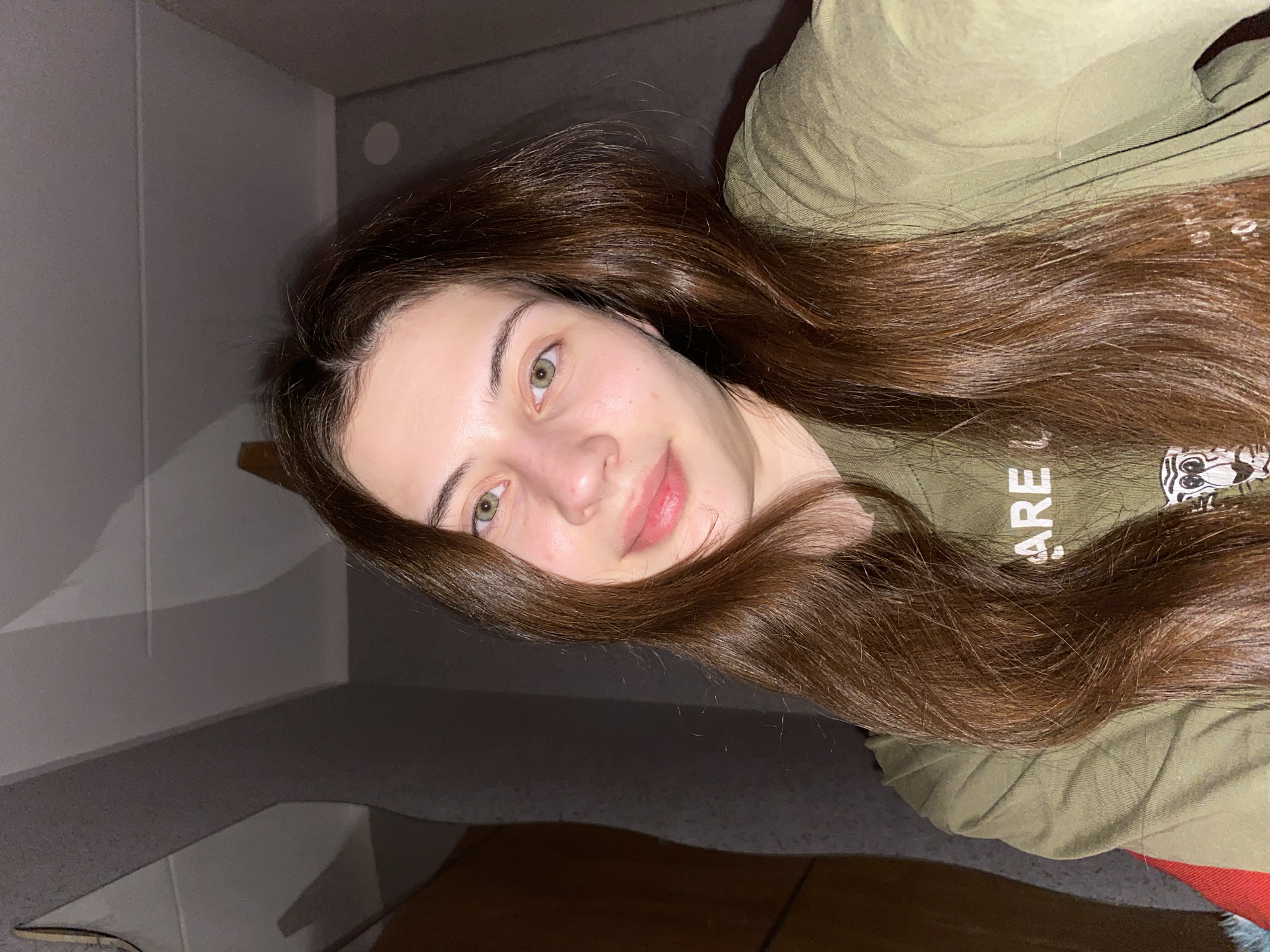 A young woman with long brown hair is lying down, partially covering an olive green t-shirt. Her face is angled towards the camera, revealing fair skin and green eyes. The background suggests an indoor setting with gray walls. No specific details indicate the location is Votkinsk, Russia.
