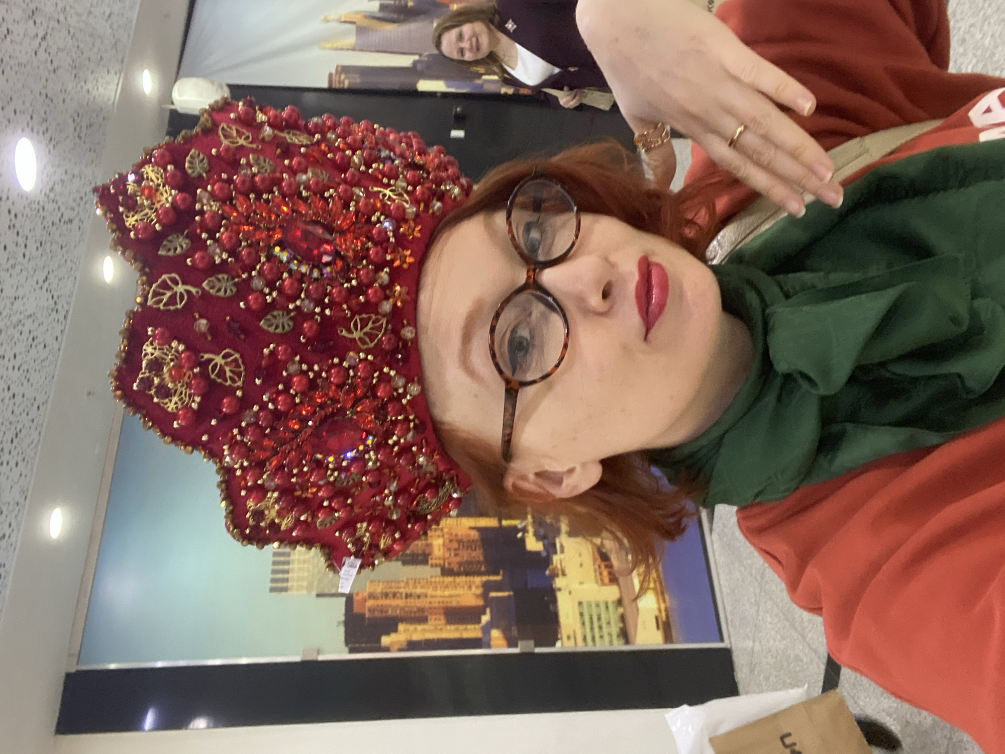 The image is a selfie-style portrait featuring a woman in the foreground, facing the camera. She has reddish-brown hair and wears round, tortoiseshell-patterned glasses. Her lips are accentuated with red lipstick, and she has a calm expression. A prominent, intricately designed red headpiece, resembling a traditional Russian kokoshnik, sits on her head. It is lavishly decorated with a multitude of red beads, sparkling jewels, and gold-colored leaf motifs. She is dressed in a green scarf and a reddish-orange top or hoodie. A gold ring is visible on her left hand, which is raised near her face.
In the soft-focused background, another woman with brown hair can be seen smiling, wearing a dark blazer over a white top. The indoor setting features a textured white wall with embedded circular lights. A large mural or poster on the wall depicts an abstract urban landscape, characterized by yellow, gold, and blue architectural shapes. A dark, horizontal partition runs below the mural. At the bottom right, a white plastic bag and a brown paper bag are partially visible, with letters "U" and "L" on the latter. The presence of the traditional headwear, likely a kokoshnik, suggests a cultural event or display, consistent with the provided location context of Yekaterinburg, Russia.