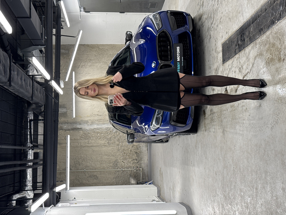 A young blonde woman stands frontally in what appears to be a luxury car showroom or detailing bay, located at Tiffany Motors, Al. Jerozolimskie 200, Warsaw, Poland. She is positioned slightly to the right of the frame's center, looking down at a light-colored smartphone held in her left hand. A small black lavalier microphone is clipped to the low V-neck of her black, long-sleeved, short blazer-style top. She wears sheer black stockings with visible garter belt straps and back seams, paired with black pointed-toe high heels. Behind her is a dark blue BMW M8 Grand Coupe, facing the viewer. The car's front license plate holder displays "TIFFANY MOTORS" with additional smaller text below it. The environment features a light gray concrete floor with visible tire marks and a metal drainage grate in the foreground. The walls are composed of raw concrete on the right and a lighter, possibly painted or paneled surface on the left, where a vertical light fixture or panel is visible. The ceiling is exposed black with industrial piping, ventilation ducts, and multiple horizontal fluorescent light fixtures.