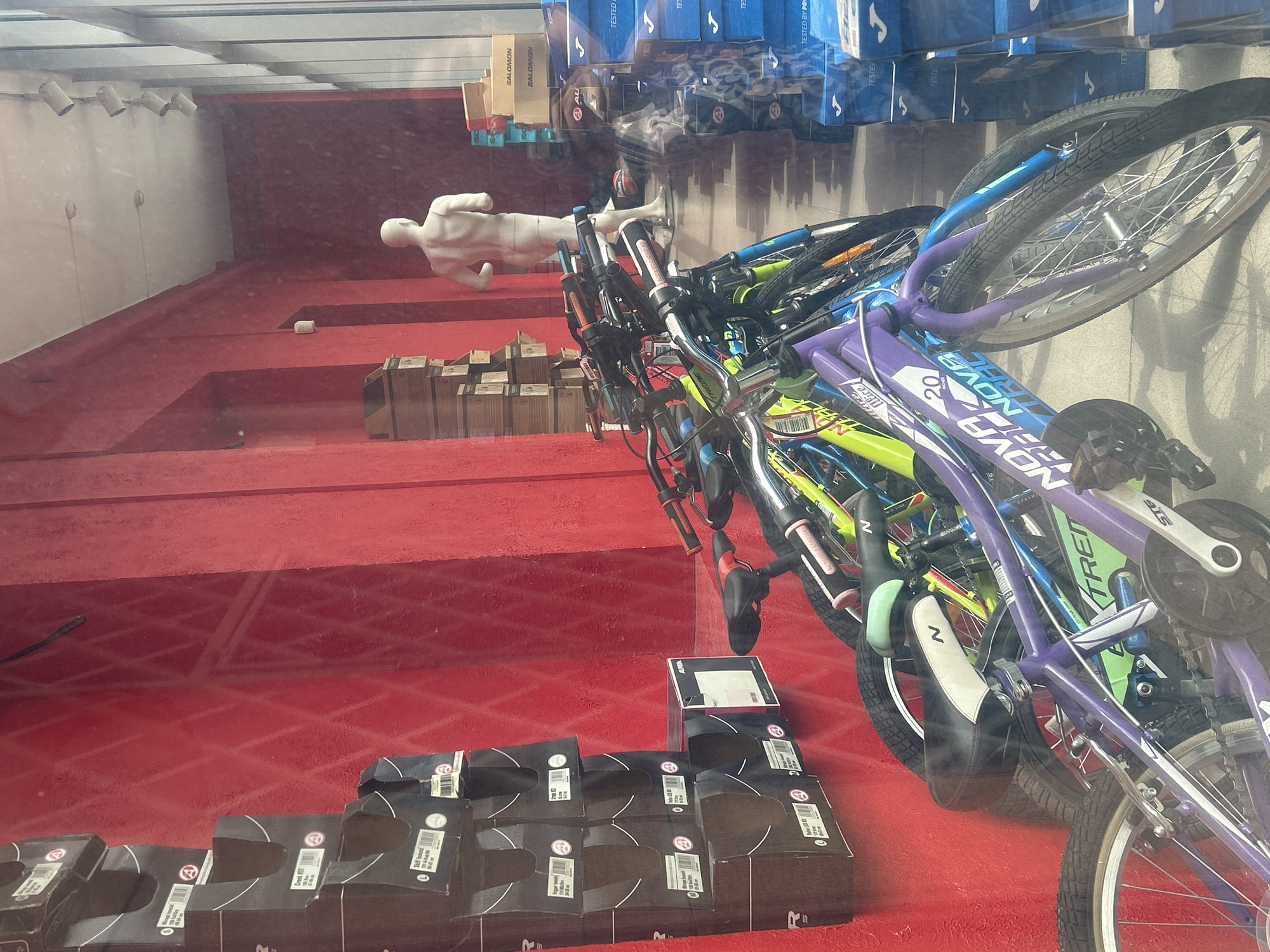 This image depicts the interior of a sporting goods store in Almaty, Kazakhstan, as seen through a window. The store has a prominent red wall with shelves displaying stacked boxes, some of which appear to contain bicycles. Several bicycles are positioned in the foreground, with their handlebars and frames clearly visible. The bicycles are of various colors, including purple and lime green. A white mannequin, resembling a male torso, stands further inside the store. The environment appears to be well-lit, with a large window on the right side reflecting the scene. Boxes with "SALOMON" branding are visible on shelves in the background, along with stacks of blue boxes that might contain shoes. The overall impression is that of a retail space showcasing sporting equipment.