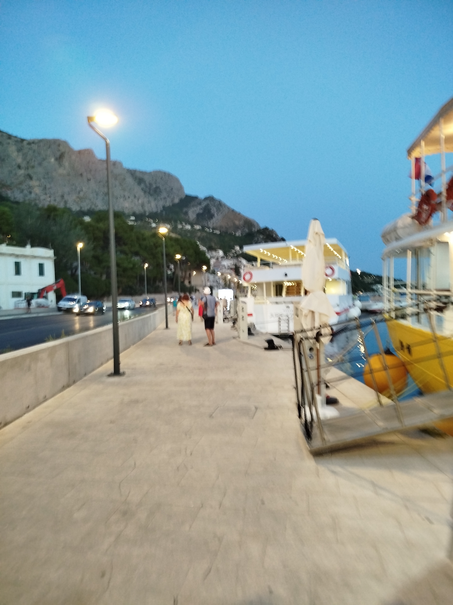 The image depicts a waterfront promenade in Omiš, Croatia, at dusk. A paved walkway runs parallel to a road with cars and a white building on the left. Beyond the road, a tree-covered hillside rises. Several lampposts illuminate the scene.
In the foreground, two individuals are walking along the promenade. A woman in a light-colored dress is ahead, followed by a man in a dark shirt and shorts.
To the right of the promenade, docked boats are visible. One boat is white with red accents and a yellow hull, featuring a metal gangway. Another larger white boat with a yellow awning and illuminated windows is moored alongside. The water reflects the lights from the boats and the sky. In the background, the lights of the town of Omiš are scattered across the hillside. The sky is a clear, deep blue, characteristic of twilight.