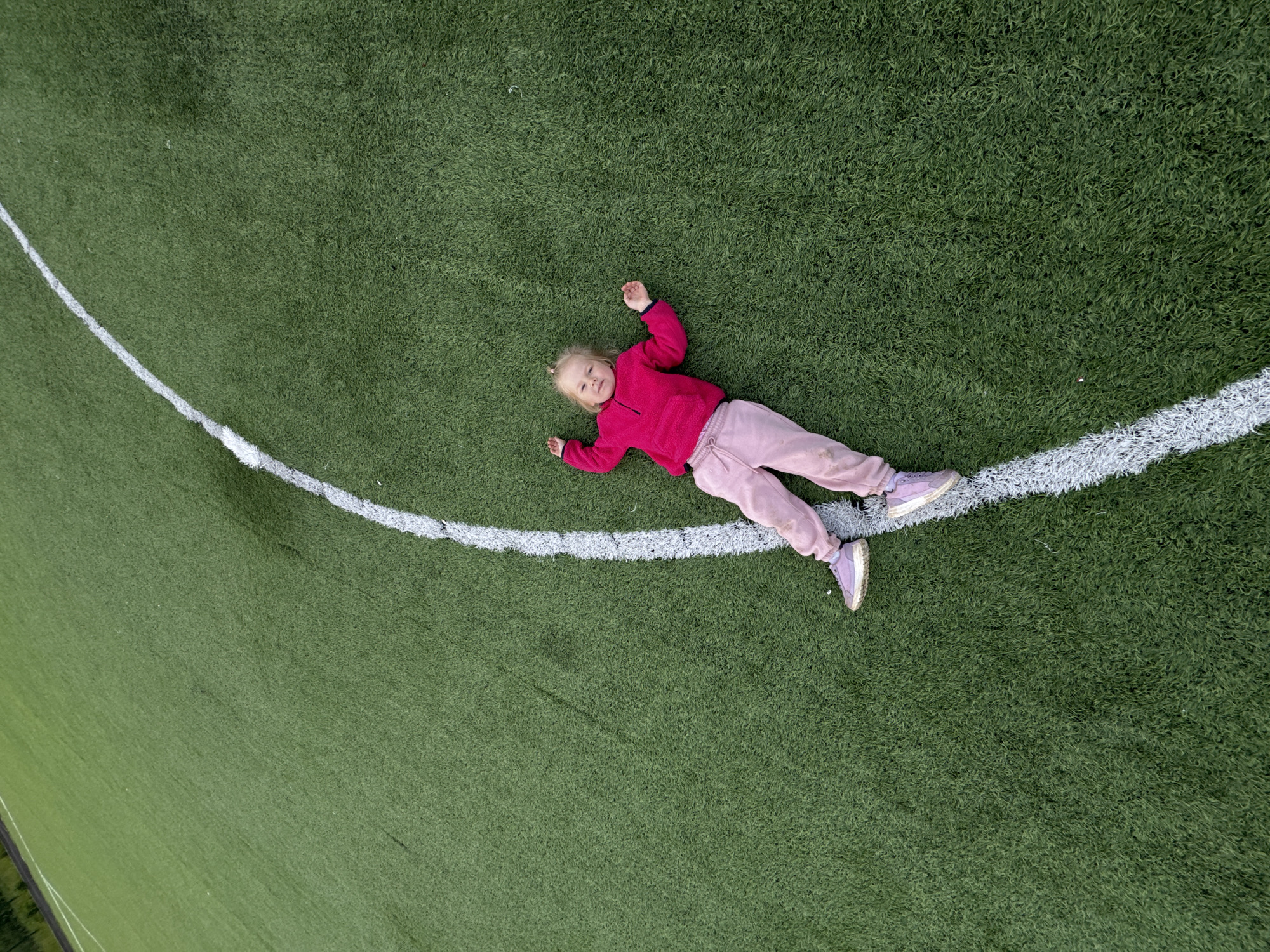 A high-angle photograph depicts a young, light-haired child lying supine on a green artificial turf sports field in Ферма, Russia. The child is dressed in a fuchsia long-sleeved top, light pink track pants, and light purple athletic shoes. She is positioned diagonally across a prominent, curved white line painted on the turf. Her arms are slightly raised with open hands, and her legs are extended. The child looks upwards towards the camera with a neutral expression. The texture of the synthetic turf is visible across the entire frame, with a dark, indistinct boundary present in the upper left background.