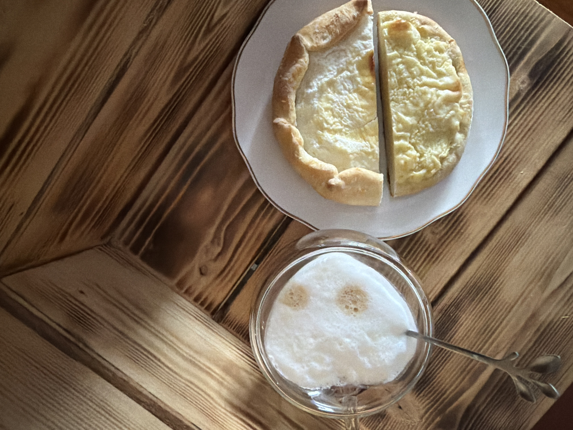 A top-down shot shows a plate with a halved cheese pastry next to a glass of white foamy liquid on a wooden table. A metal spoon is partially submerged in the glass. The wood has a textured grain pattern. The image was captured in the city of Chaikovsky, Russia.