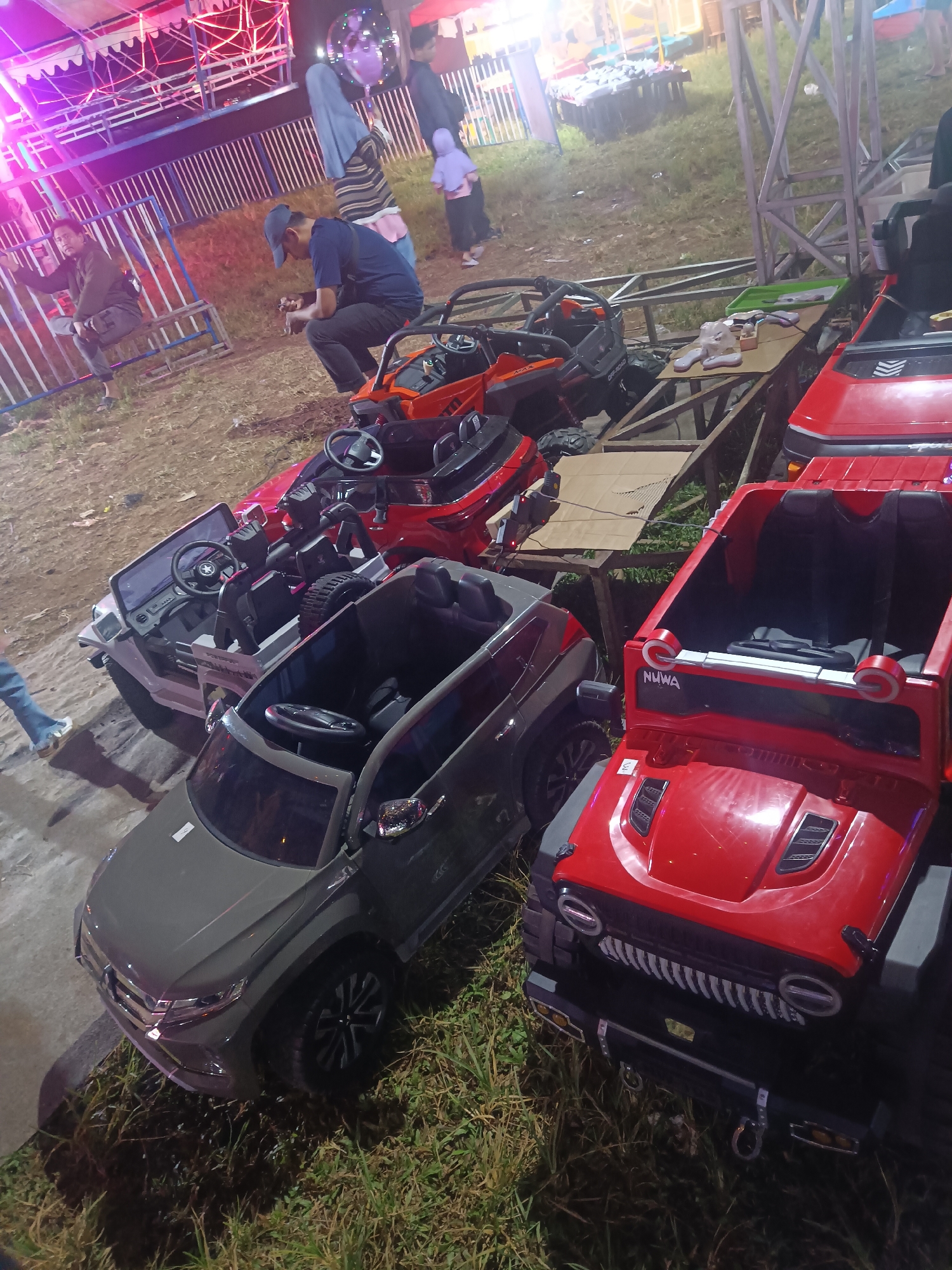 The image shows a scene at what appears to be a night market or amusement area, likely in Depok, Indonesia. Several children's ride-on electric toy cars are parked in rows. The cars are mostly jeeps and SUVs, in colors like red, gray, and dark orange. Some have visible steering wheels and seats. One red jeep has the word "NUWA" visible on its front.
In the background, there are hints of amusement park rides with colorful lights, suggesting it's nighttime and the area is festive. A few people are visible in the background, including adults and children, some looking towards the rides or walking around. One man in a blue shirt and a cap is sitting down, looking at something in his hands, perhaps a phone. Another man is seen sitting on a bench with a railing. People are dressed casually, some in t-shirts and pants, others in traditional clothing.
The ground appears to be a mix of dirt and grass. The lighting suggests it is evening or night, with artificial lights from the attractions illuminating the scene. There are no clear indicators of the weather, but the overall atmosphere seems relaxed and social, typical of an evening out at an entertainment venue.