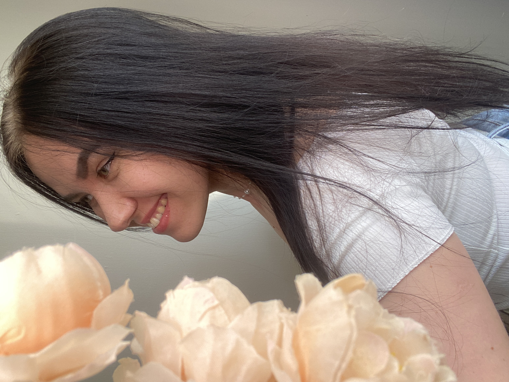 An image of a young woman with long, dark hair, seen in profile, is displayed. She is looking upwards and to the left, with a slight smile on her face. She is wearing a white, ribbed top. To the left of the woman, a cluster of large, pale peach-colored flowers fills the foreground, partially obscuring her face and shoulder. The background is a plain, light gray wall. The woman is likely indoors, as indicated by the lighting and the backdrop. No specific location within Chaikovsky, Russia, can be determined from the visual information.