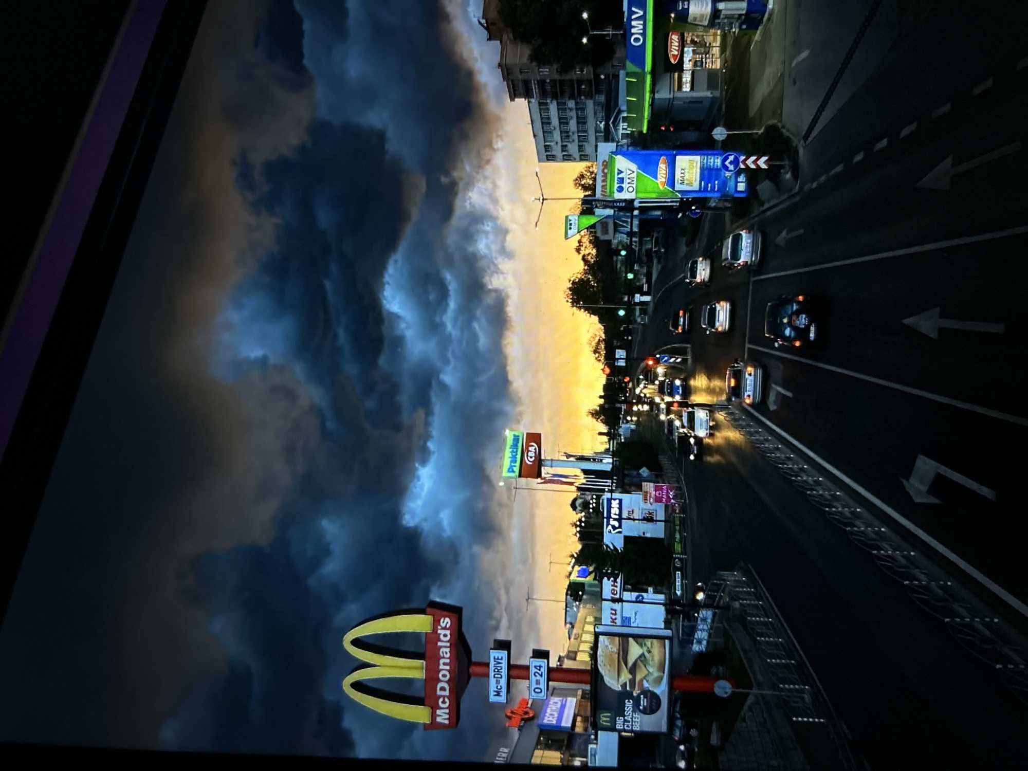 An outdoor, low-light photograph captures an urban commercial street scene at dusk or early evening, characterized by heavy, dark storm clouds overhead and a striking orange-yellow sunset on the horizon. The upper sky is dominated by turbulent, deep blue-grey and black clouds, with a brilliant horizontal band of orange and yellow light from the setting sun visible between the clouds and the cityscape.
A multi-lane asphalt road, appearing wet and highly reflective, occupies the midground and foreground, with numerous vehicles in motion. White painted arrows on the road indicate straight-ahead and right-turn lanes, and a median barrier with white posts and railings separates traffic directions. Streetlights are illuminated, casting elongated reflections on the wet surface.
On the left side of the road, a prominent, brightly lit McDonald's pylon sign features the iconic yellow "M" logo and "McDonald's" text, along with "McDrive" and "0-24" (24 hours) indicators. Below this, a large billboard displays an advertisement for a "BIG CLASSIC BEEF" burger. Signs for "Decathlon" and other businesses like "KUK", "ISK", and "Mania" are partially visible further back, set against commercial buildings.
On the right side of the road, an OMV gas station is clearly visible with its distinctive green and blue branding, displaying "OMV" and "VIVA MAXX Motion" signs. Further down, a tall pylon sign advertises "Практикер" (Praktiker, a home improvement store) with "CBA" (a supermarket chain) displayed below it, both in Cyrillic script. Distant apartment buildings and trees complete the background.
Numerous vehicles, including sedans and SUVs, are present on the road, with headlights and taillights visible and reflecting. The overall scene suggests a major thoroughfare or boulevard in a commercial zone of an Eastern European city, specifically identified as **Sofia, Bulgaria**, due to the combination of retail chains (OMV, Praktiker, CBA, Decathlon) and the use of Cyrillic script.