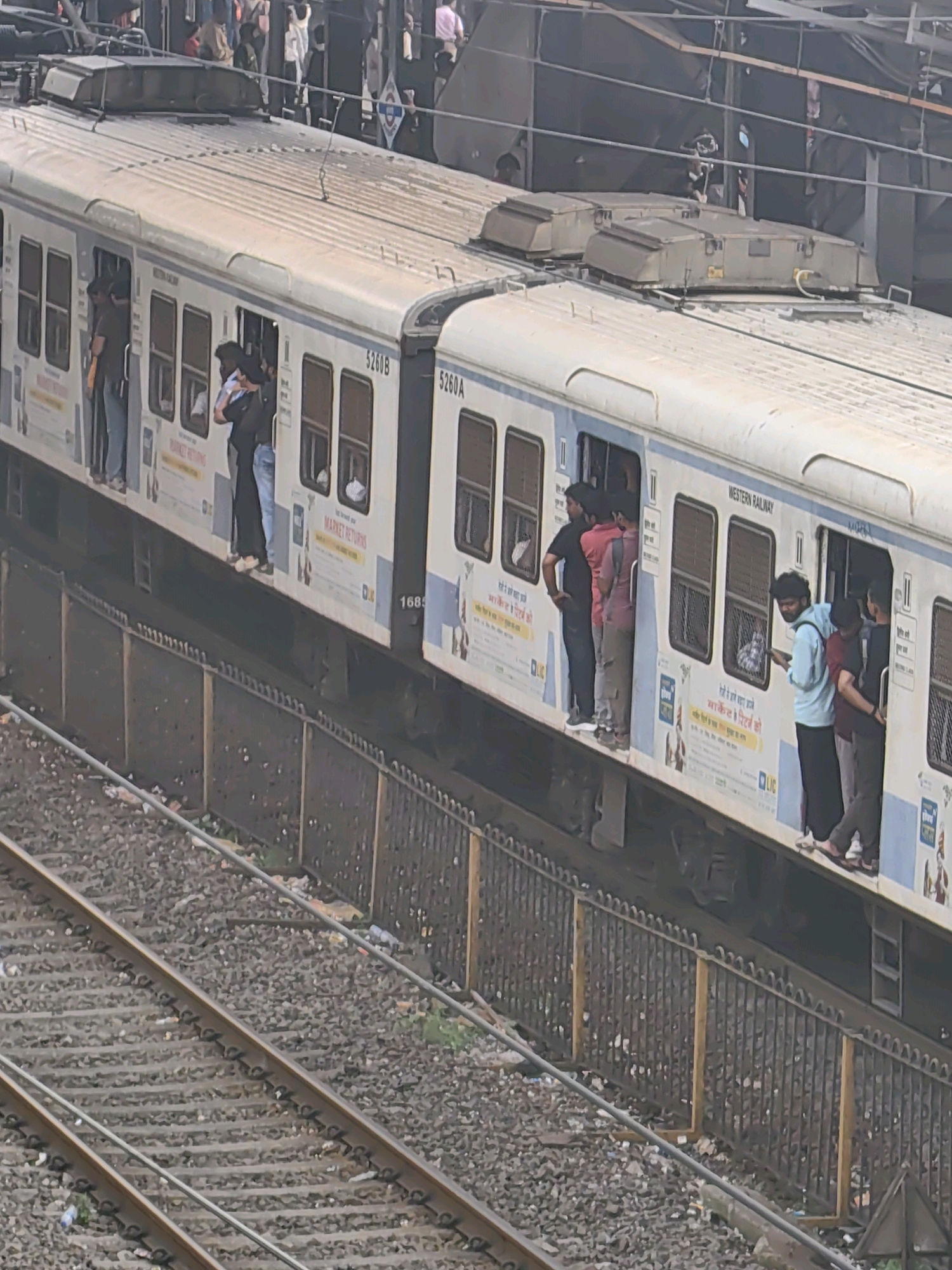 The image depicts a crowded Mumbai local train, likely at a station, with passengers standing both inside and hanging out of the open doors. The train carriages are white with blue and yellow trim, and the words "WESTERN RAILWAY" are visible on one carriage. Several individuals are seen in the doorways, with some leaning out. The train is positioned on tracks adjacent to a platform, which is indicated by a metal fence. The ground around the tracks is littered with debris. In the background, other passengers and structures are partially visible, suggesting a busy urban environment characteristic of Mumbai.