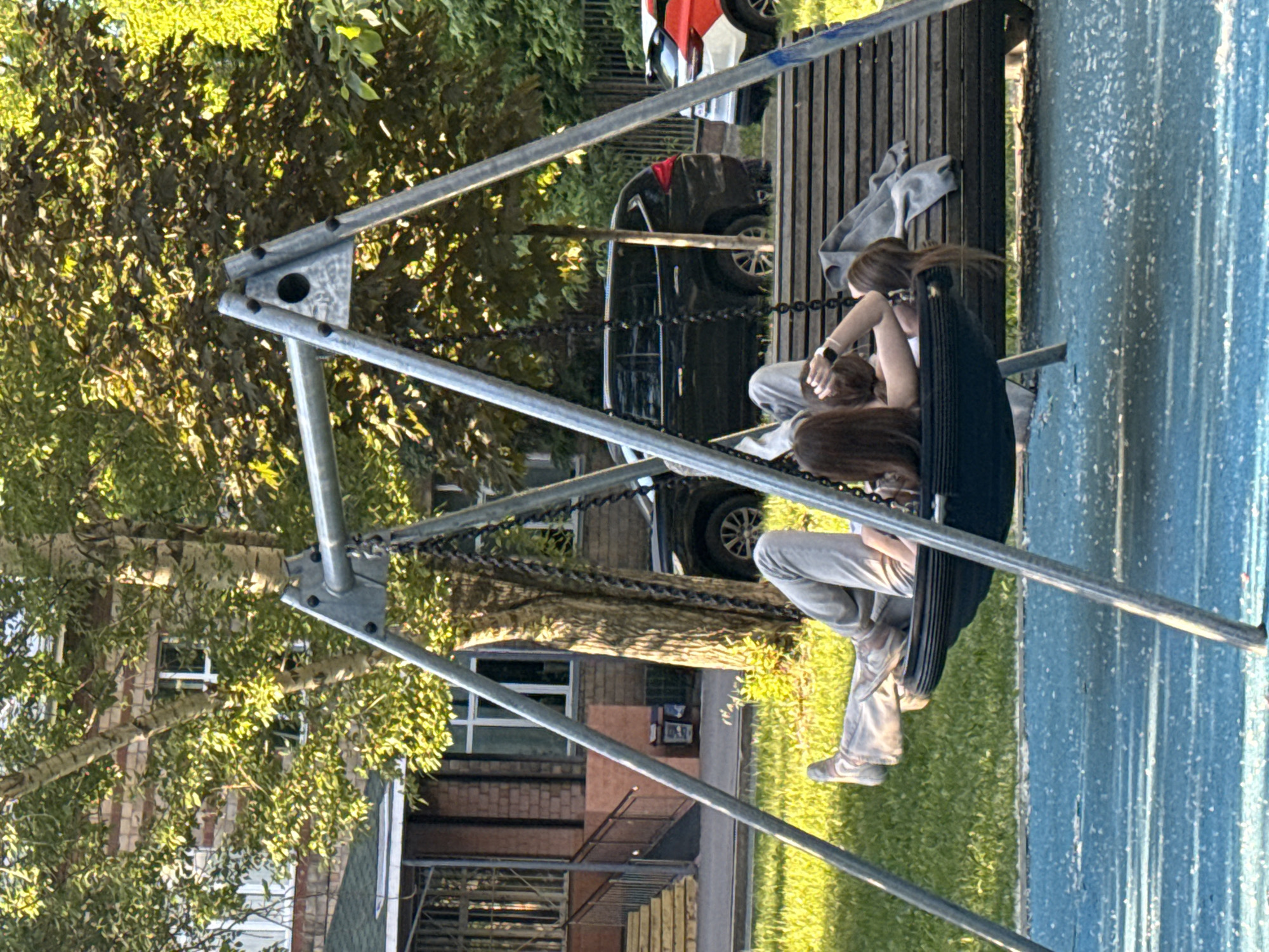 The image depicts two individuals, likely young women, seated on a tire swing outdoors. The swing features a metal A-frame support structure and a black tire as its seat. The person closer to the viewer is wearing light-colored pants and sneakers, with long brown hair. Another person is seated behind her on the same swing, also with long hair and wearing a light top. They appear to be relaxed, possibly conversing or resting.
Behind the swing, a wooden slatted platform or bench is visible, with a light grey piece of clothing draped over it. In the mid-ground, a dark sedan is parked, and another car with white and red elements is partially visible. The background is dominated by lush green trees and foliage, suggesting a natural, park-like environment. Several brick buildings with windows can be glimpsed through the trees.
The ground cover includes green grass on the left side of the frame and a textured, light blue surface, possibly a rubber playground floor or sports court, on the right. The scene is brightly lit by natural sunlight, casting shadows, which indicates a sunny day, likely in the late afternoon. No visible text is present in the image. The setting appears to be an urban or suburban recreational area, consistent with a park or playground in Moscow, Russia.