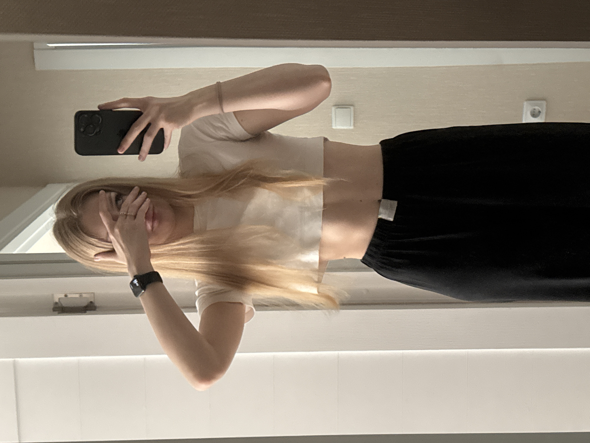 A young Caucasian female with blonde hair, wearing a white crop top and black pants, stands in front of a mirror in what appears to be a residential interior. She is holding a black smartphone in her right hand, taking a selfie, and covering her eyes with her left hand. The background shows a wall with a light switch and an electrical outlet, suggesting a typical home environment. The overall scene depicts a casual moment of self-photography. The location is not identifiable as the city of Perm, Russia.