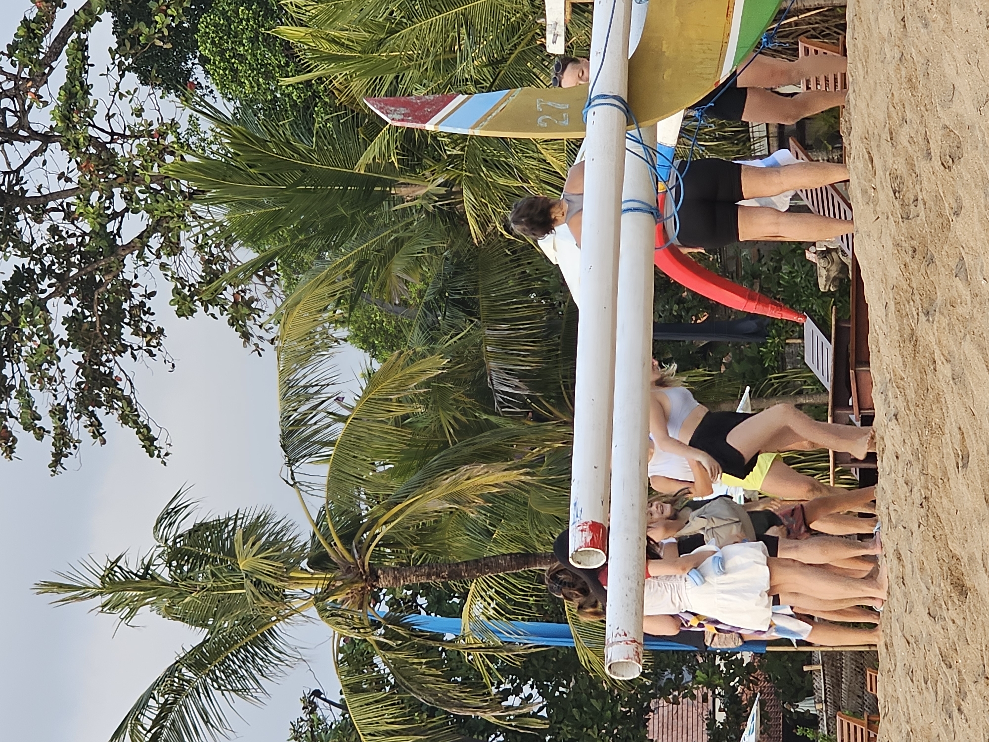 In Denpasar, Indonesia, a group of young women are gathered on a sandy beach near a traditional outrigger boat. The boat, partially visible on the right, is adorned with a colorful fin featuring the number "27". The women, dressed in casual beach attire, appear to be interacting with the boat, with some holding onto white poles that extend from it. Palm trees and lush vegetation surround the scene, suggesting a tropical coastal environment. The sand is textured and uneven, with some wooden lounge chairs visible in the background. The lighting indicates it is daytime, with diffused light suggesting overcast conditions or late afternoon.